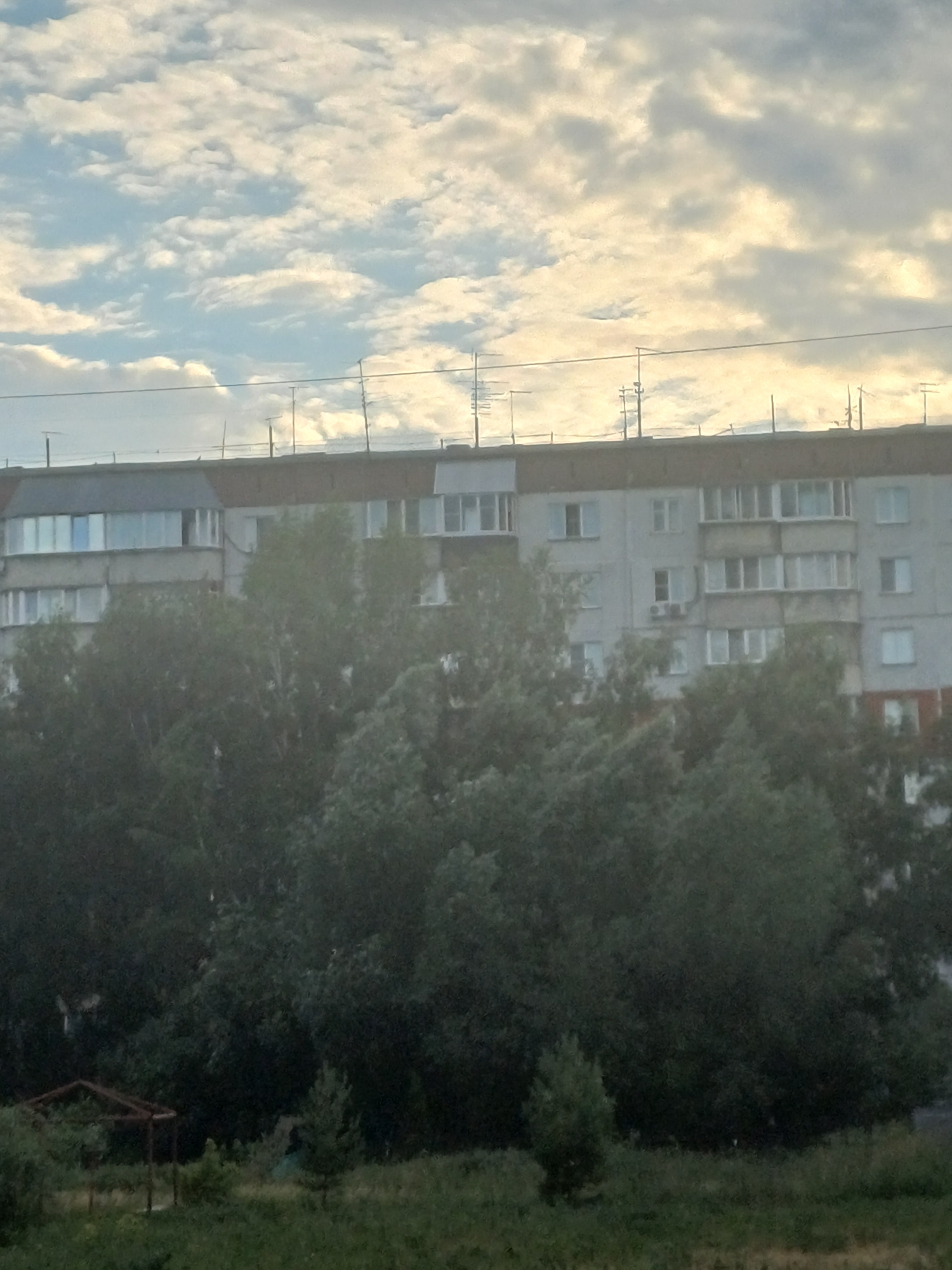 This media file is a photograph taken in Novosibirsk, Russia. The image depicts a building with multiple stories, likely an apartment complex, adjacent to a lush green area with trees and grass. The building's facade is light-colored, with visible windows and balconies. There are power lines running vertically along the left side of the image, in front of a sky filled with scattered, bright clouds. The lighting suggests it is daytime, possibly late afternoon or early morning, given the soft, diffused light illuminating the clouds. The overall atmosphere is calm and residential. There is no text visible in the image.