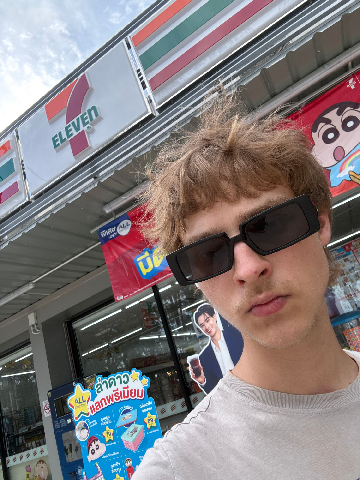 This is a portrait-oriented, medium-angle, eye-level shot taken outdoors, likely during the day with overcast skies. A young person with blonde, tousled hair and wearing black, rectangular sunglasses is the central subject, looking towards the camera. They are wearing a plain white t-shirt.
The setting appears to be in front of a 7-Eleven convenience store, as indicated by the prominent "7 ELEVEN" logo visible on signage and the store's characteristic colorful exterior. The store's interior is visible through the windows, with shelves stocked with merchandise and fluorescent lighting. Various promotional posters and displays are also visible, some featuring cartoon characters. One poster, written in Thai, advertises a "PREMIUM COLLECTION" with stars and cartoon characters, mentioning "ALL member" and prices in Thai Baht. Another poster shows a cartoon character that appears to be from the anime "Crayon Shin-chan." A small, blue flag with a yellow logo and Thai text is also hanging outside the store. The overall impression is casual and mundane, with the subject posing for a photo in front of a commercial establishment. The lighting suggests it's daytime, but the overcast sky doesn't provide harsh shadows.