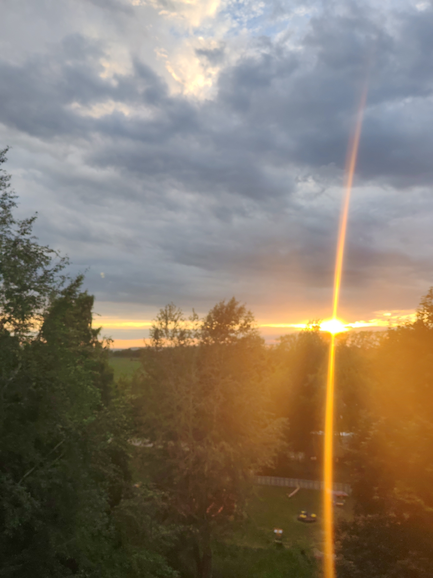 A sunset is captured with dramatic lighting through a dense cloud cover over a rural or suburban landscape. The sun, a bright white sphere, is partially obscured by a band of orange and yellow clouds along the horizon. A strong, vertical sunbeam, emanating from the sun, pierces the clouds, casting a bright, golden glow. The foreground is dominated by dark green trees, appearing somewhat out of focus, which frame the view of the sky and the distant landscape. The lower portion of the image reveals a grassy area with some small, indistinct structures, possibly part of a backyard or small park. The overall impression is one of natural beauty and tranquility during the twilight hours. The image is taken from a slightly elevated perspective, looking out over the trees towards the horizon.