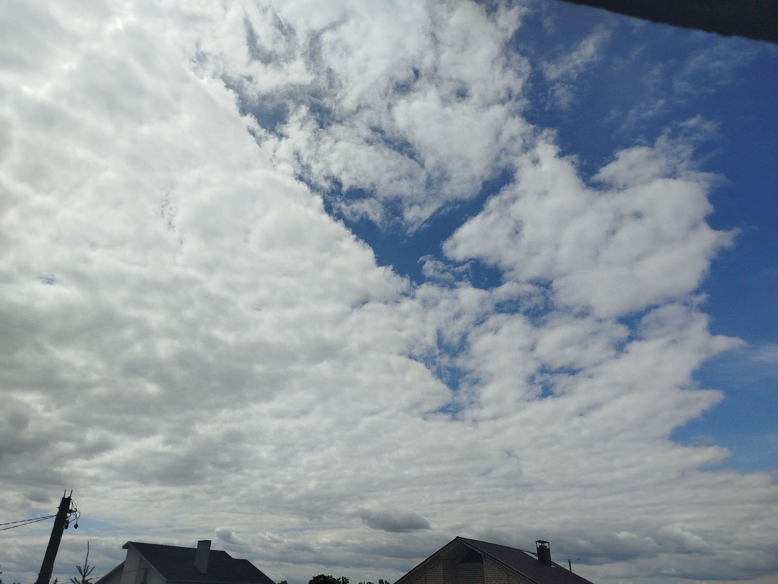 A wide-angle, slightly low-angle shot captures a cloudy sky over a semi-rural landscape in Kapyĺ, Belarus. The sky is a prominent feature, with a mix of heavy, overcast clouds on the left and brighter, scattered cumulus clouds against a vibrant blue sky on the right. A dark, angled object, likely part of a building's roof or an overhang, frames the top of the image.
In the lower portion of the frame, glimpses of rooftops and a utility pole suggest a residential or semi-developed area. The utility pole on the far left is dark and angled, with wires extending from it. Two house rooftops are visible: one on the left with a dark roof and angled sides, and another on the right with a similar dark roof, a visible chimney, and brickwork. The presence of trees and greenery is hinted at in the distance, between and beyond the houses.
The scene appears to be set during the daytime, with the quality of light and cloud formations indicating it could be late morning or afternoon. The weather is mixed, with significant cloud cover but patches of clear blue sky, suggesting changeable conditions. There is no text visible in the image. The overall mood is one of an ordinary day, characterized by the dynamic sky.