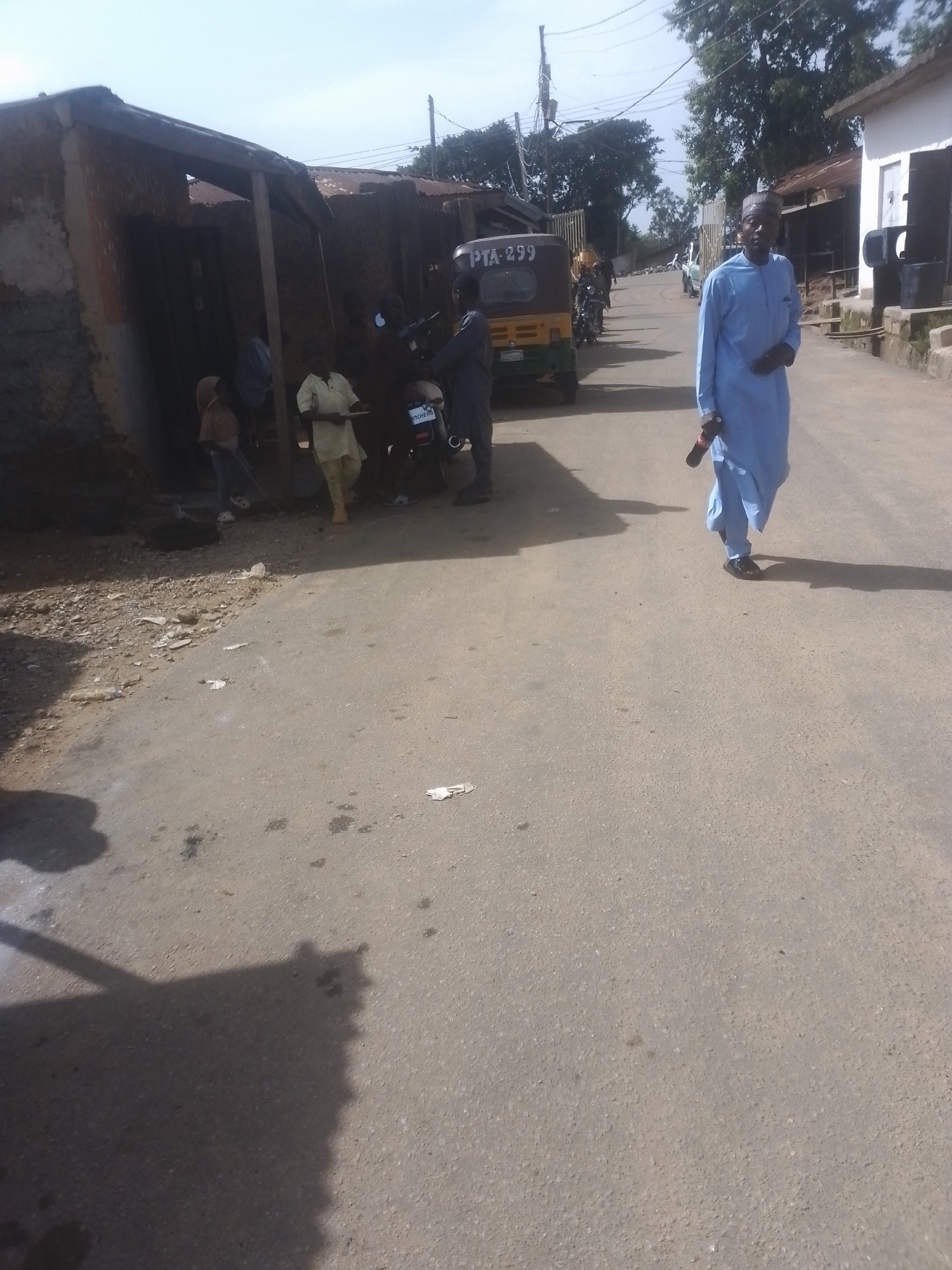A medium shot captures an outdoor scene with several people and objects on a paved road. A man in a light blue robe and a cap walks towards the viewer, holding a dark object in his right hand. Beside him, on the left side of the road, are several people gathered near some buildings. One person appears to be standing near a motorcycle. Further down the road, a three-wheeled vehicle, possibly a tuk-tuk, is parked. The buildings appear to be made of brick and corrugated metal, suggesting a simple, perhaps rural or semi-urban setting. The sky is visible and appears to be clear and bright, indicating daytime. The road itself is asphalt, with some debris and dark stains scattered on its surface. The overall impression is of a casual moment in a community. There is some visible text on the back of the three-wheeled vehicle: "PTA - 299".