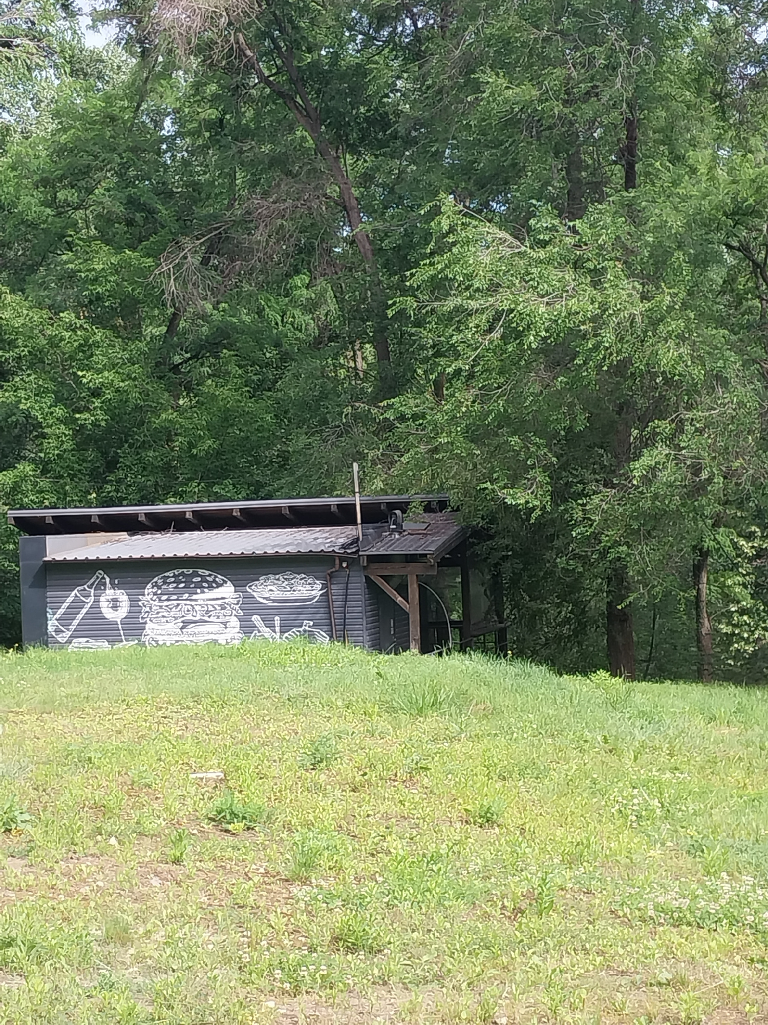 A low-angle, eye-level shot captures a small, dark-colored building with a corrugated metal roof, nestled amongst lush green trees and overgrown grass. The building features a mural on its side, depicted in white outline style, showcasing a large hamburger, a bottle of drink, a glass, and a plate of food. The scene appears to be in an outdoor, natural setting.