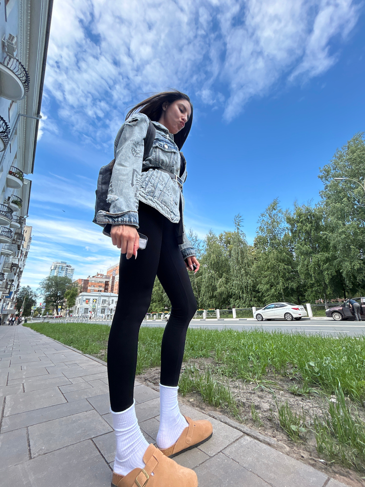 A low-angle, full-body shot captures a young woman standing on a paved sidewalk. She is wearing a light-wash denim jacket with a graphic print, black leggings, white crew socks, and tan slip-on shoes. Her dark hair is down, and she is holding a smartphone in her right hand. She appears to be making a kissy face, looking up towards the camera.
The background reveals a vibrant scene. A bright blue sky with scattered white clouds dominates the upper portion of the image. Below the sky, a lush green tree line runs horizontally, separating the sky from a city street. Cars are visible on the street, including a white sedan and a darker vehicle. A white fence separates the grassy area from the street. In the distance, urban buildings with multiple stories can be seen.
The paving stones of the sidewalk are laid in a repeating pattern. The overall lighting suggests it is daytime, likely a sunny afternoon, given the clear sky and shadows. The setting appears to be an urban environment in Novokuybyshevsk, Russia. The woman's relaxed posture and facial expression suggest a casual, possibly playful moment. No visible text is present in the image.