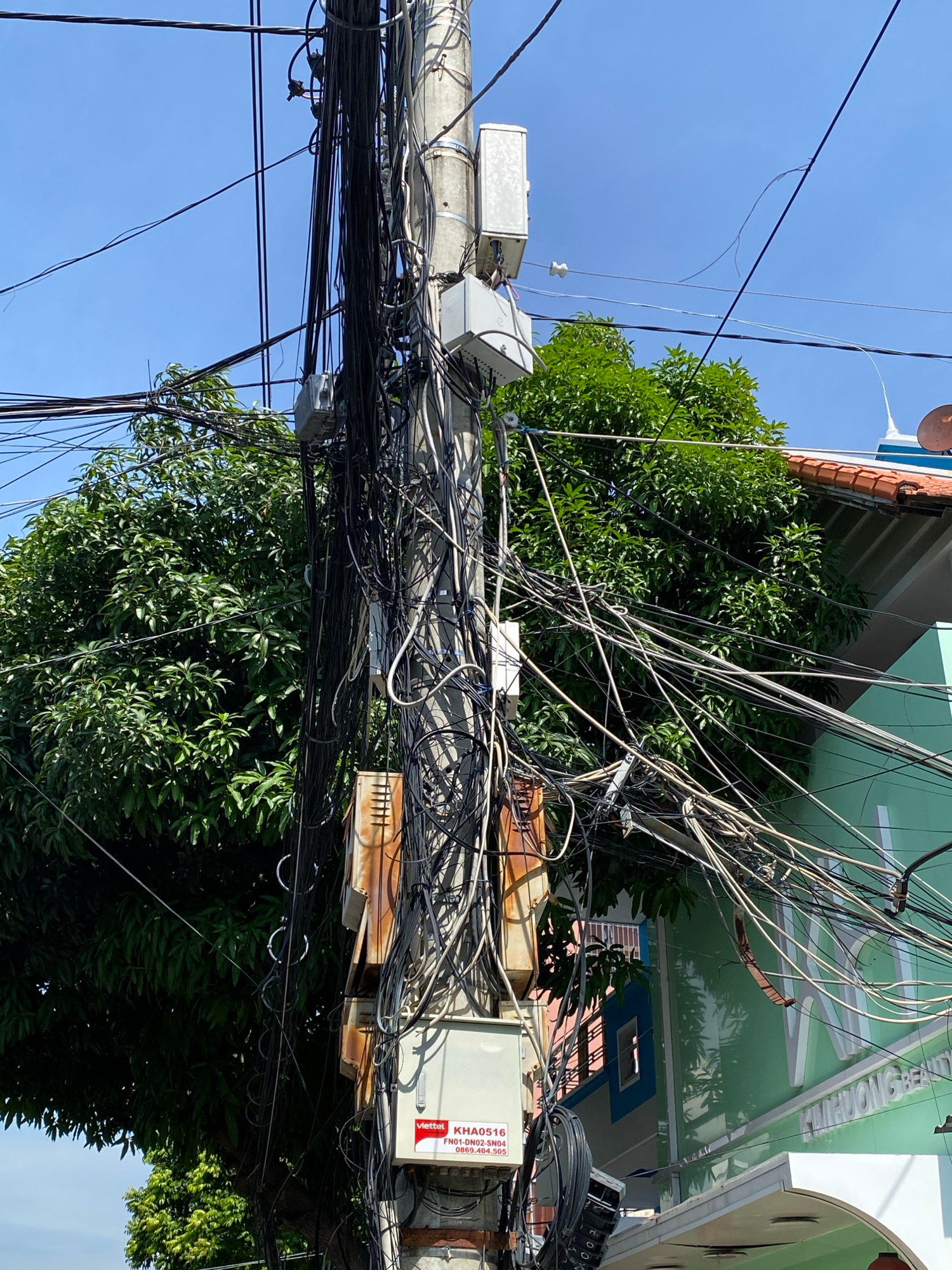 The image captures a concrete utility pole densely covered with a chaotic tangle of black and white electrical wires, likely in an urban setting in Nha Trang Ward, Vietnam, given the signage on the building. A small white electrical box with "Viettel KHA0516" and a phone number printed on it is attached to the pole. Two white rectangular boxes, possibly telecommunications equipment, are mounted higher up on the pole. Behind the pole, a large, vibrant green tree with dense foliage is visible, its leaves illuminated by sunlight. To the right of the pole and tree, a building with a light green facade and an orange-tiled roof is partially visible. The building has a sign with white lettering, though the full text is not discernible. The sky above is clear and blue. The overall scene depicts a common urban landscape element in Vietnam, characterized by the extensive and somewhat disorganized network of overhead utility wires.