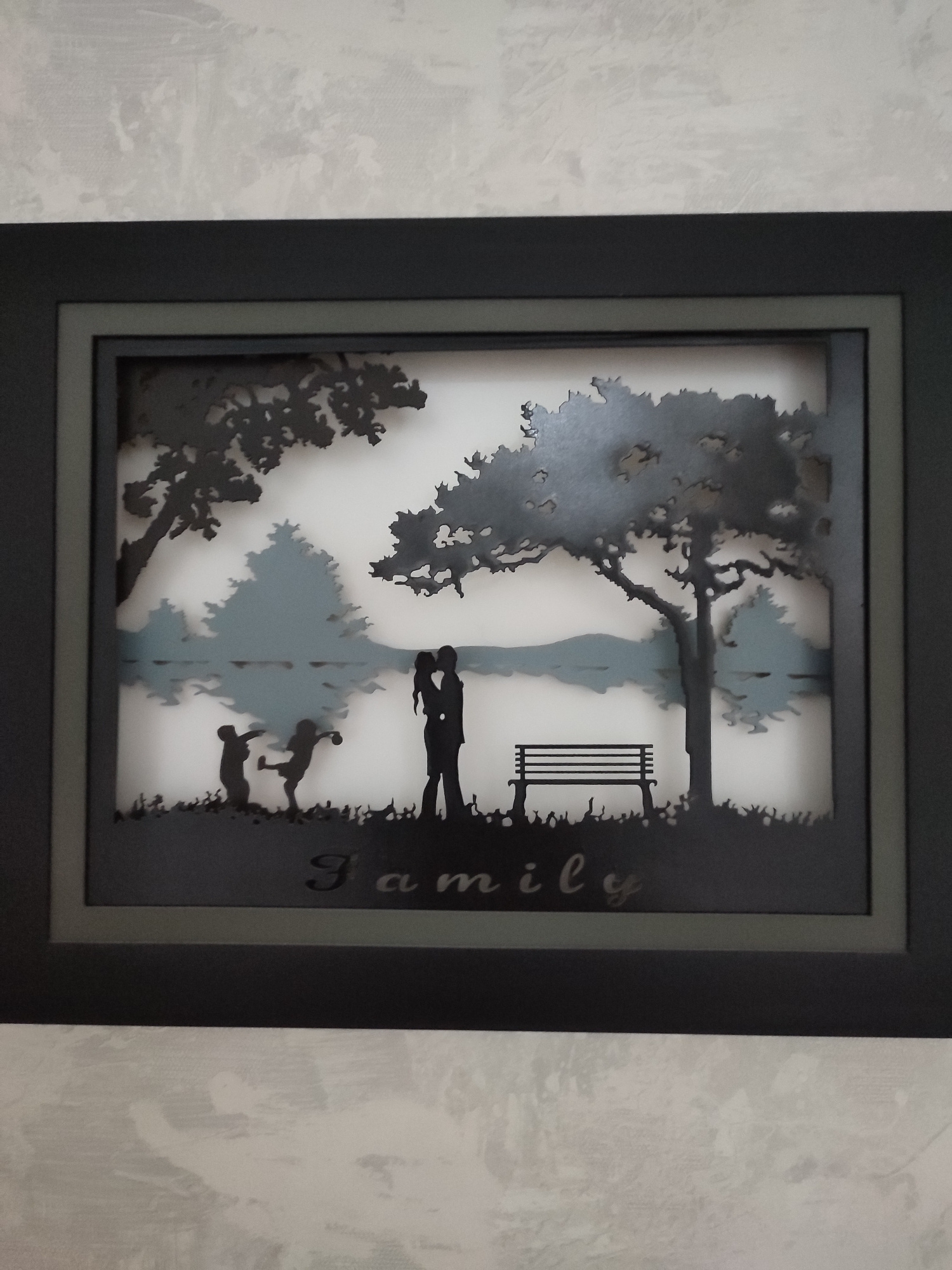 This is a framed shadow box artwork featuring a silhouette scene. The foreground depicts a grassy area with a bench on the right and a couple embracing on the left, with two children playing in front of them. The middle ground shows a body of water with trees reflected on its surface. The background consists of a landscape with trees and a body of water. The overall scene is peaceful and evokes a sense of family togetherness. The artwork is framed in a dark rectangular frame. The word "family" is inscribed in script below the figures. The lighting suggests it is daytime.