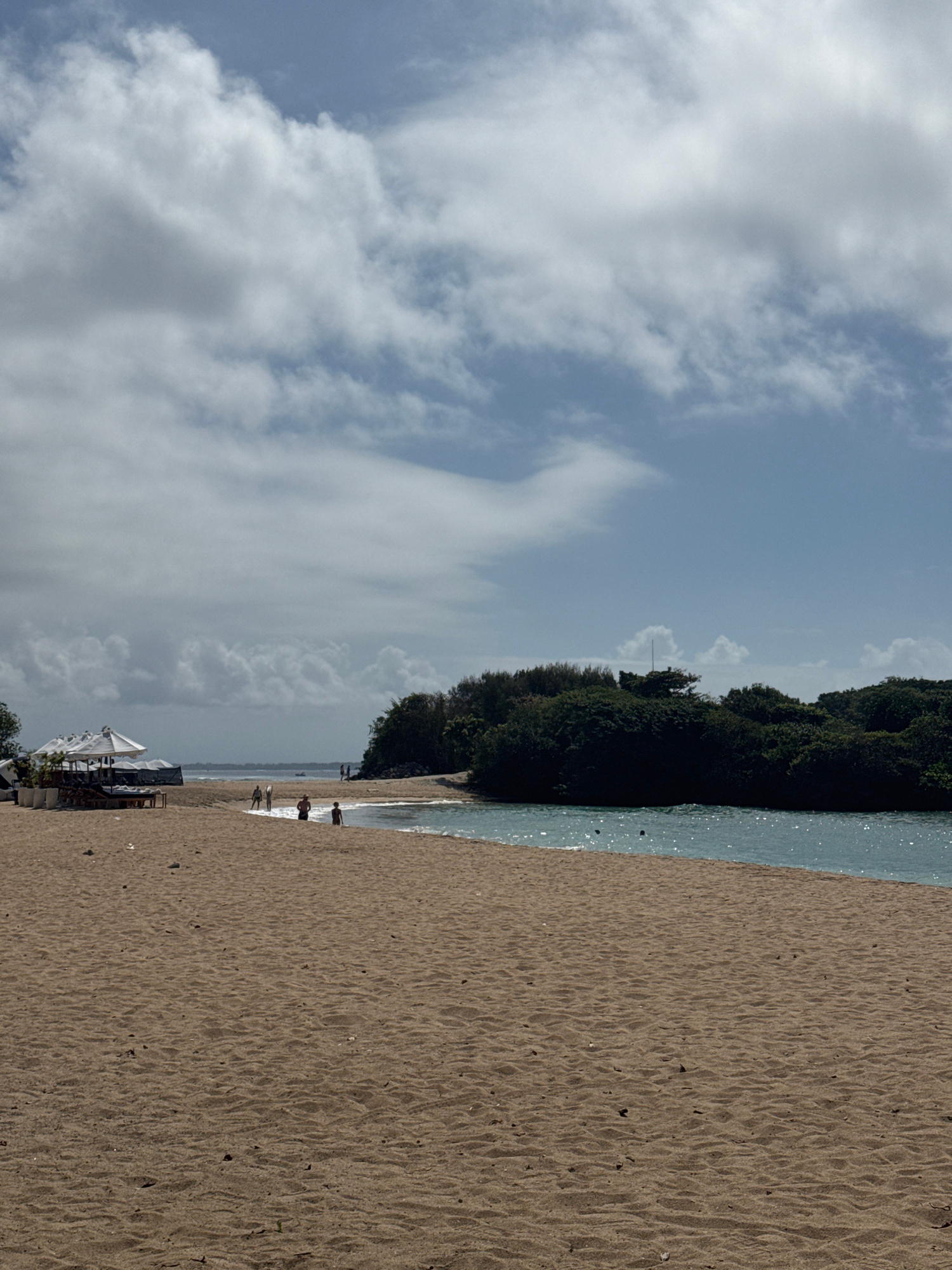 A sandy beach features several lounge chairs and umbrellas in the foreground, set against a backdrop of lush green trees and a body of water. Several people are scattered across the beach and near the water's edge. The sky above is partly cloudy, with patches of blue visible.