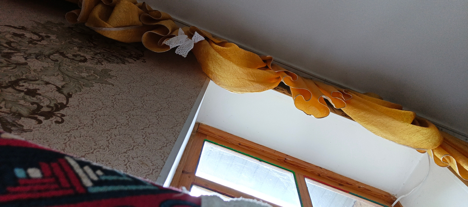 The image shows an interior view looking upwards towards a window and a decorative yellow valance. The valance is textured and has ruffled edges, adorned with a small white lace bow. The wall to the left features ornate, patterned wallpaper in shades of beige and brown with green accents. To the right, a wooden window frame is visible, with some evidence of wear. The window glass appears somewhat dusty. A portion of a patterned rug or fabric with red, blue, and white geometric designs is in the lower left foreground. The ceiling is a plain white. This scene is located in a room in Kök-Jangak City, Kyrgyzstan.