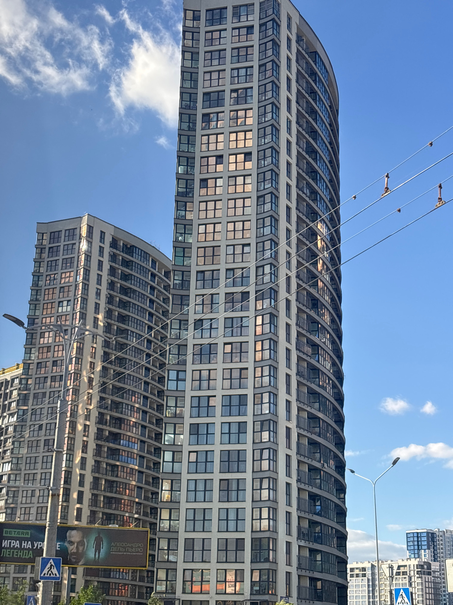 The image shows a street view in the Senitsky Selsoviet, Belarus, featuring several modern high-rise residential buildings. The primary focus is on two prominent, curved apartment buildings with numerous windows and balconies, reflecting the sky and surrounding environment. To the left, a billboard is visible, displaying advertisements with text in Cyrillic and images of people. A lamppost and traffic signs, including a pedestrian crossing sign, are present on the street level. Overhead, power lines traverse the frame, supported by insulators. The sky is blue with scattered white clouds. In the background, other buildings and structures are discernible, indicating an urban or suburban setting. The lighting suggests it is daytime, possibly late afternoon due to the warm reflections on the building facades.