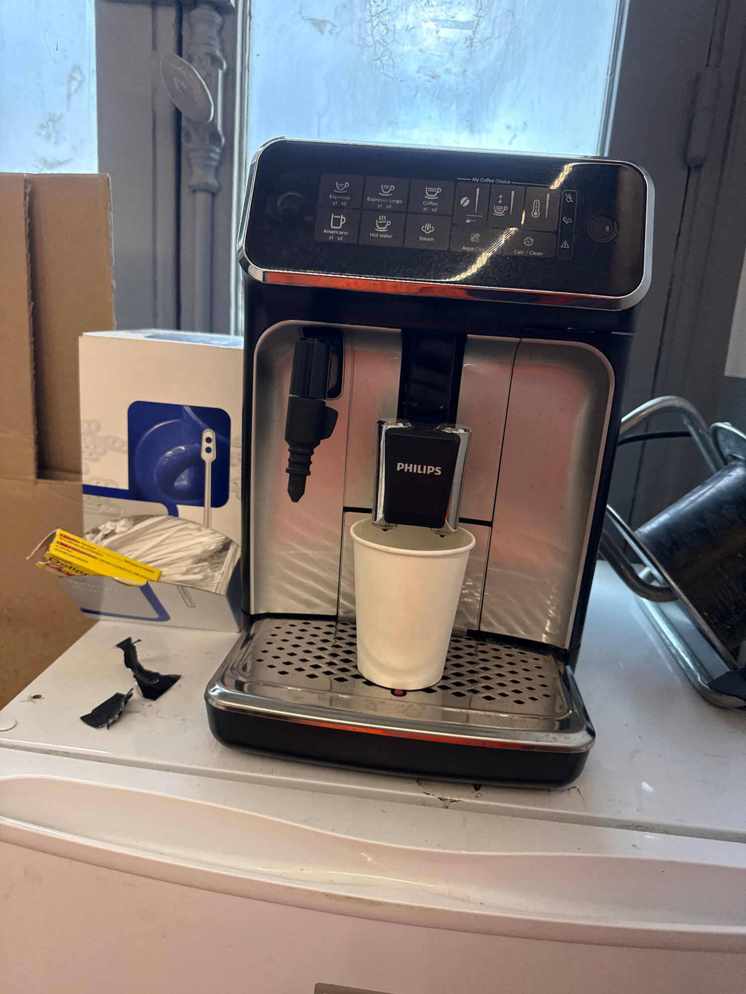 A Philips automatic coffee machine is positioned on a white countertop. A white paper cup is placed beneath the dispensing spout of the machine, ready to receive coffee. To the left of the coffee machine, a white box containing coffee stirrers is visible. In the background, a window with distressed panes is present, and to the left of that, stacks of cardboard boxes are observed. The scene appears to be indoors. There is no indication that this location is in Paris, France.
