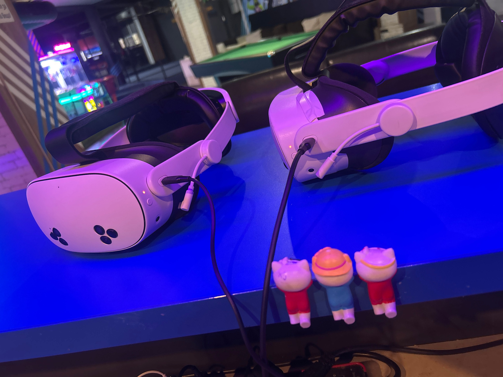 The image displays two white virtual reality headsets positioned on a bright blue surface. The headsets have black headbands and are connected by wires. In the foreground, three small figurines are lined up: one wearing red, one in blue with an orange hat, and another in red. The background suggests an entertainment venue, with a pool table visible and colorful neon lights in the distance, hinting at an arcade or gaming area. The lighting in the scene is predominantly purple and blue, creating a futuristic or gaming atmosphere. There is no visible text in the image. The overall impression is that of a modern gaming setup ready for use, likely in the evening or at night given the artificial lighting. The setting appears to be indoors, possibly a VR arcade or a dedicated gaming space.