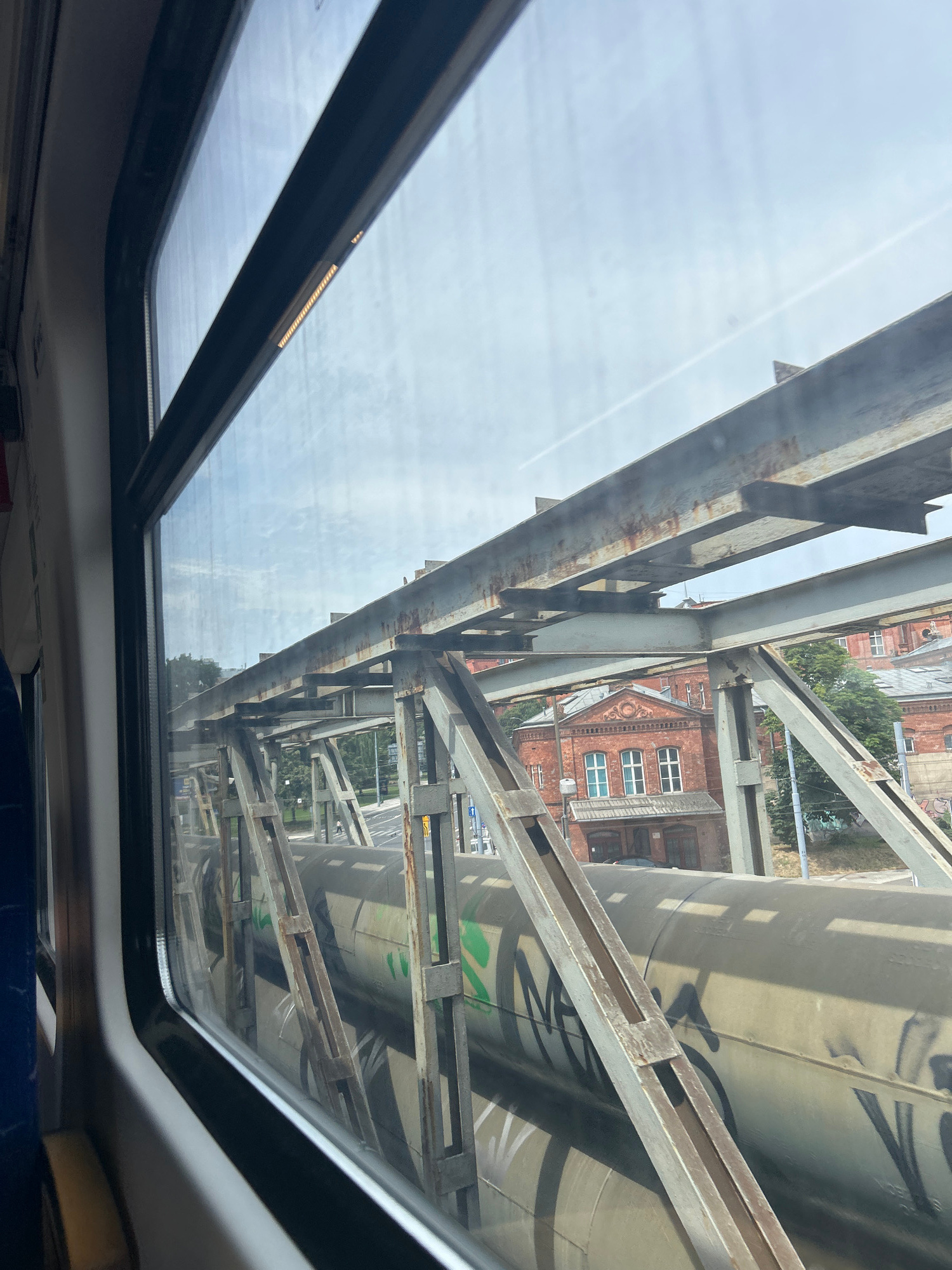 The image shows a view from a train window looking out at a concrete pipe structure that is part of the infrastructure of the city of Szczecin, Poland. The pipe is large in diameter, covered in graffiti, and supported by a metal framework. In the background, there are red brick buildings with arched windows, characteristic of industrial architecture in Szczecin. Trees and a street with a road and sidewalk are also visible. The sky is cloudy, suggesting overcast weather. The train window frame is visible in the foreground, indicating the perspective is from inside a moving vehicle.