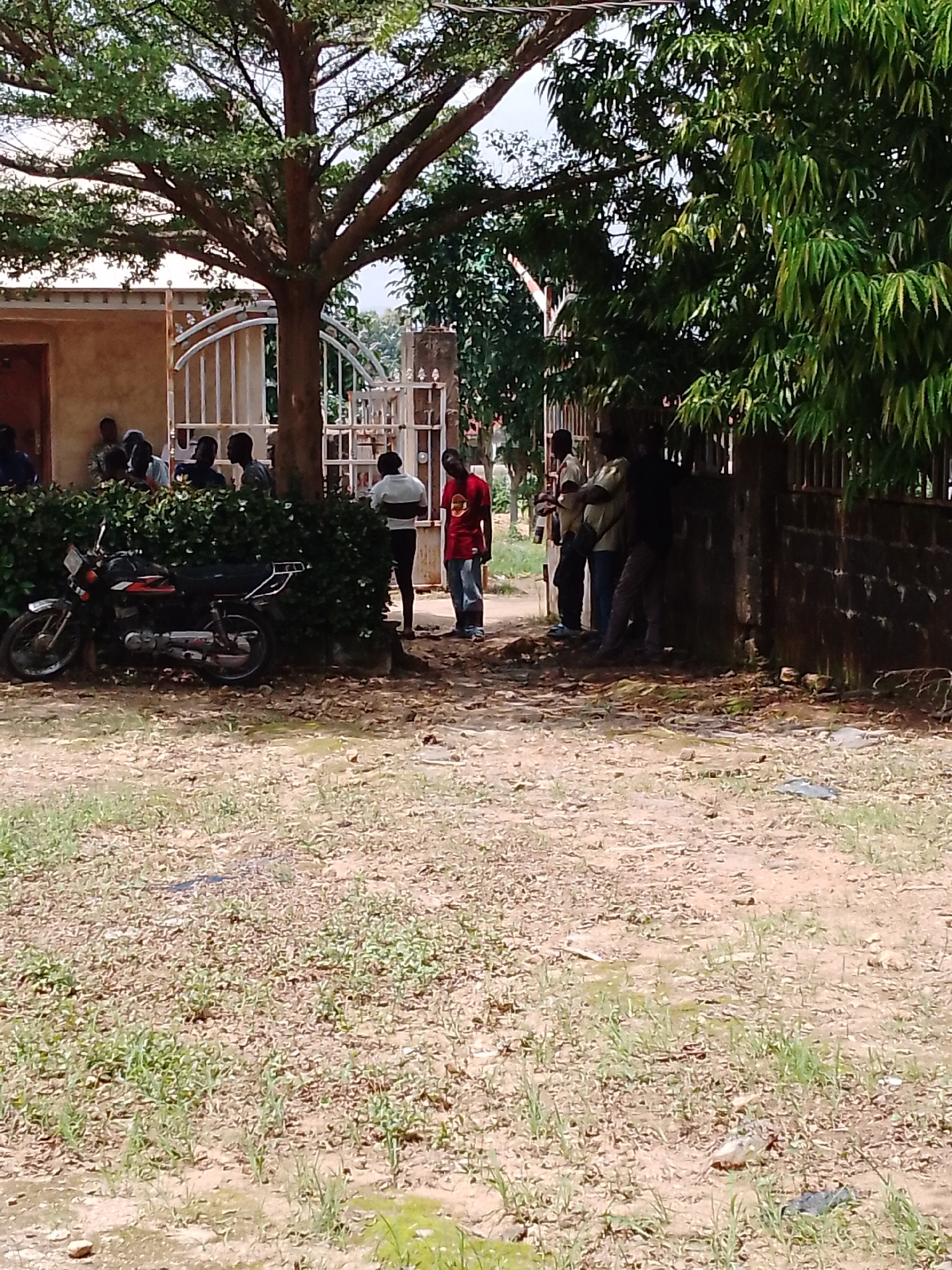 The image shows a group of people gathered near a building with a metal gate and a large tree. In the foreground, a motorcycle is parked next to a hedge. The ground is a mixture of dirt and sparse vegetation. The overall scene suggests an outdoor gathering or waiting area, possibly in a residential or community space. The time of day appears to be daytime, with ample natural light. The weather seems to be clear, indicated by the brightness of the scene and the shadows cast by the tree. The people are casually dressed and appear to be conversing or interacting in small groups. Some are standing, while others are leaning against the wall or tree. One person in a red shirt stands out from the others. The building behind them has a light-colored facade and a brown door. The gate is ornate and white. The dense foliage of the tree and the other plants create a slightly shaded environment. The location cues, such as the style of the building, the gate, and the vegetation, are consistent with a tropical or sub-tropical climate. There is no visible text in the image.