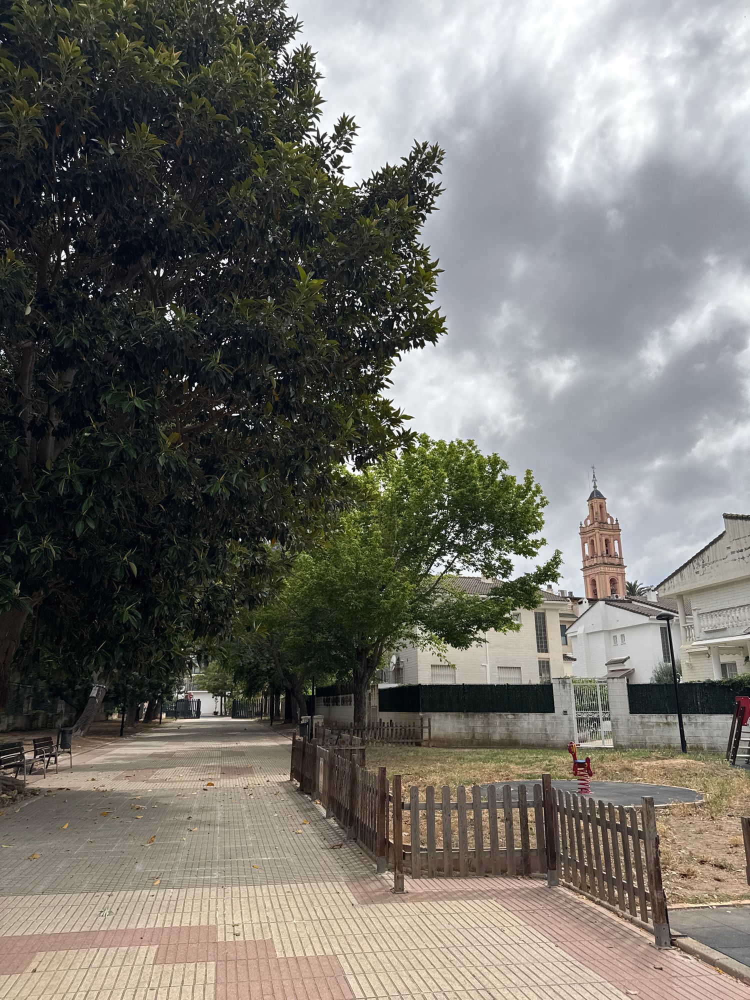 A tree-lined pathway stretches into the distance under a cloudy sky in Benirredrà, Spain. On the left, a large, mature tree with dark green leaves dominates the foreground, its branches reaching towards the overcast sky. To the right of the pathway, a small, fenced-off playground area contains a red spring rider. Beyond the playground, houses with white facades and tiled roofs are visible. In the background, a light pink bell tower with arched openings and a dark steeple rises above the buildings, indicating a church or similar structure. The pathway is paved with rectangular tiles in a checkerboard pattern of beige and red. Benches are placed along the left side of the path. The overall scene depicts a public space with residential elements and a touch of recreational area.