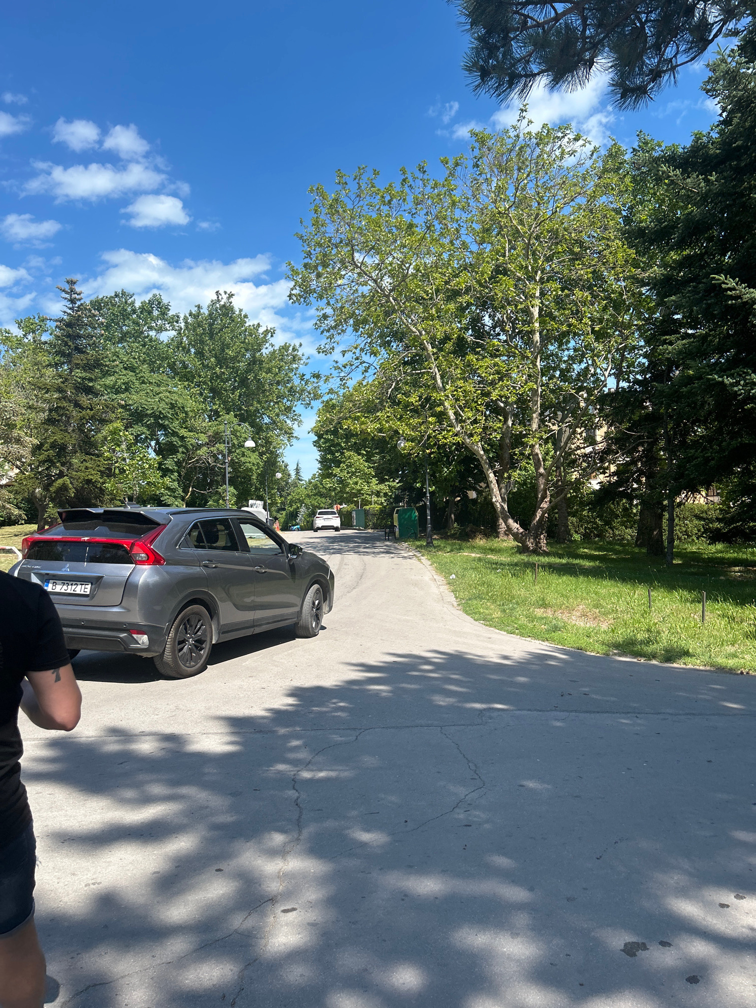 The image shows a gray Mitsubishi Eclipse Cross parked on a paved path in a park. The car is facing away from the camera and slightly to the right. The path curves to the right and is lined with green grass and trees. The sky is blue with scattered white clouds.
In the foreground, a person's arm and shoulder are visible, suggesting they are standing next to the car or taking the photo. The person appears to have a tattoo on their arm.
In the background, more trees and greenery are visible, along with a street light and a trash can. Further down the path, another white vehicle is parked. The overall scene is bright and sunny, indicating it is daytime. The presence of lush green grass and trees suggests a warm season.
The license plate of the Mitsubishi is visible and reads "B 7312 TE". The scene appears to be a public park or a recreational area in Varna, Bulgaria. The atmosphere is relaxed and peaceful, characteristic of a sunny day in a park.