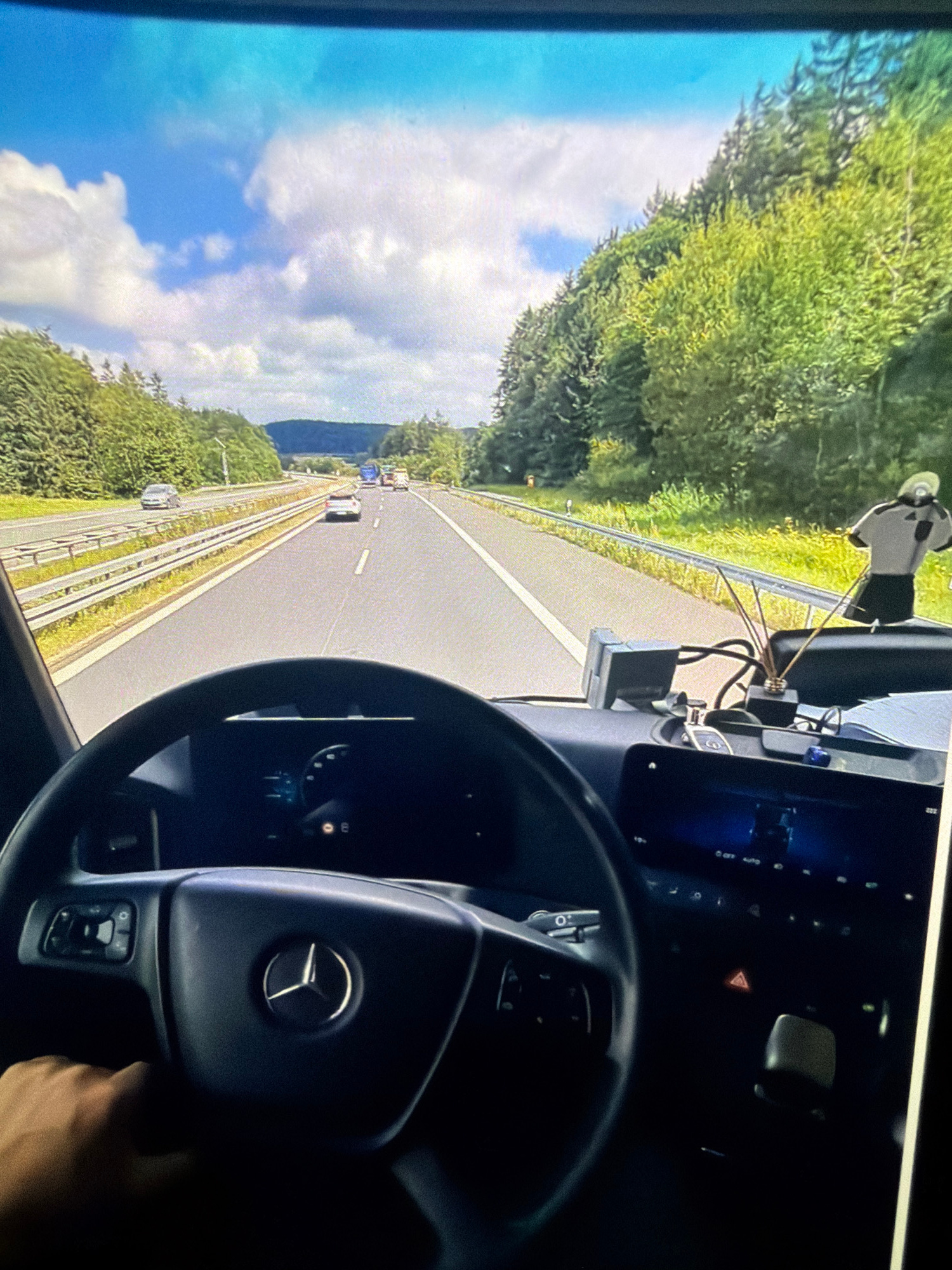 The image captures a first-person perspective from inside a vehicle, likely a truck, as it travels on a highway. The steering wheel, with a Mercedes-Benz emblem, is visible in the foreground, and a hand is resting on it. The dashboard shows a digital display and various controls. Outside the vehicle, the highway stretches ahead, flanked by lush green trees and hills. The sky is blue with scattered white clouds. Several other vehicles, including cars and trucks, are visible on the highway ahead. A decorative air freshener is placed on the dashboard. The scene appears to be a scenic drive through a rural or wooded area on a sunny day.