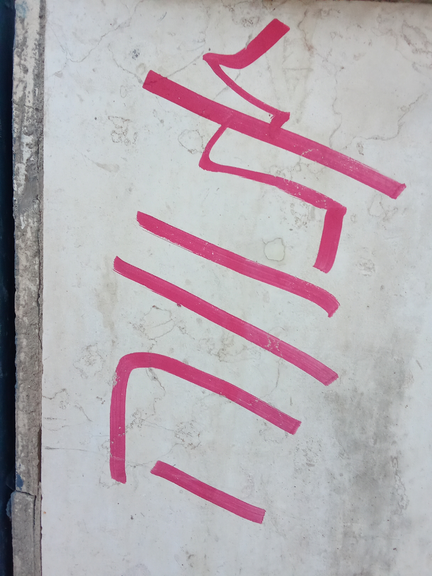 A close-up, vertically oriented shot shows red graffiti on a light-colored, textured surface that appears to be a wall or a sidewalk. The graffiti consists of thick, angular strokes of red paint. Some strokes are long and straight, while others are curved or broken. The background surface exhibits a mottled texture with faint lines and some darker smudges. The image is cropped such that a section of a darker, rougher material, possibly concrete or stone, is visible on the left edge, suggesting it is a border or edge of the surface. The overall impression is of urban decay or street art.