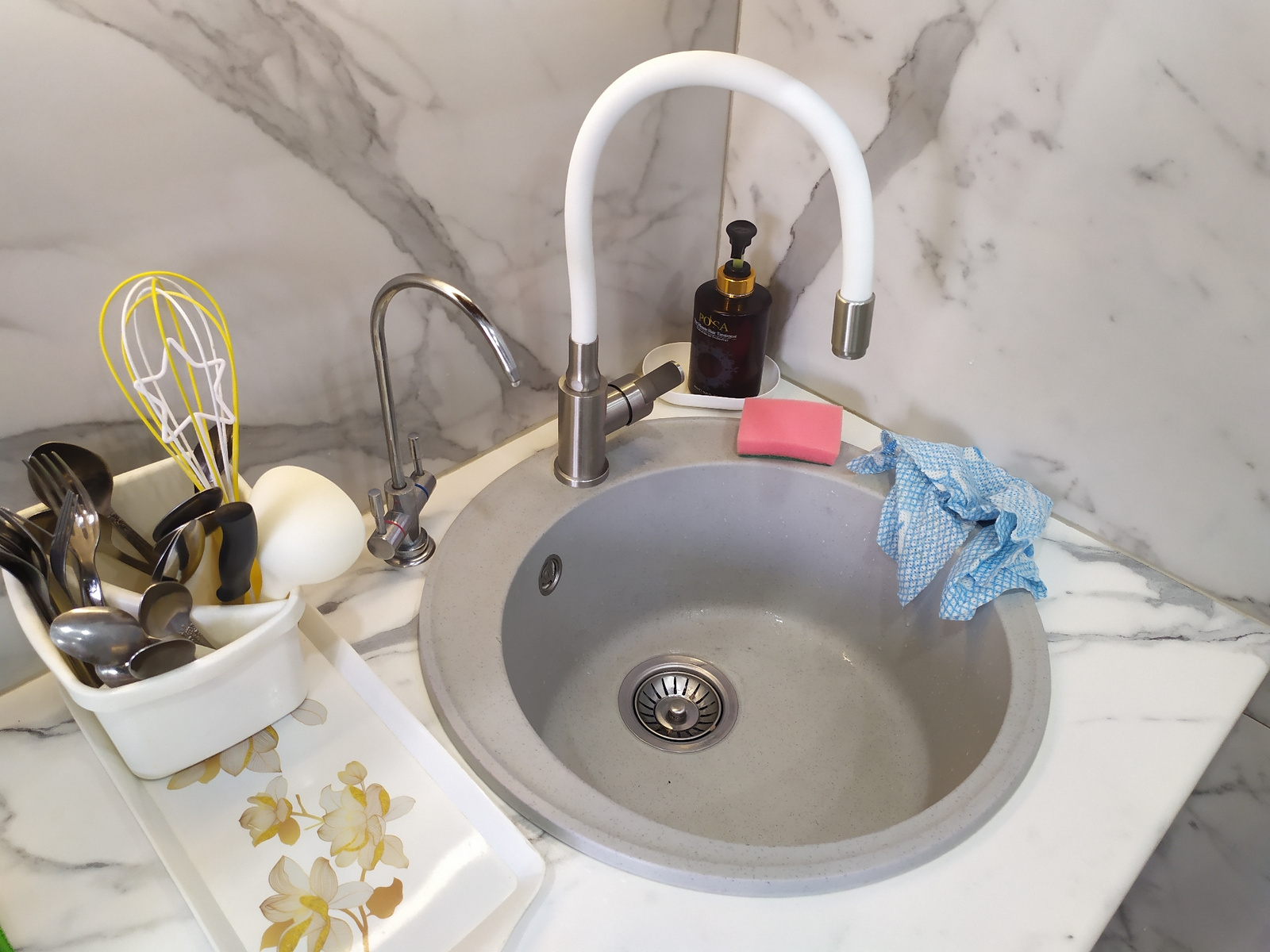 A close-up, high-angle shot shows a kitchen sink area with marble-patterned walls and countertops. The sink itself is round, light gray, and made of composite material, with a stainless steel drain. A silver faucet with a flexible white spout is installed. Next to the sink, on a white soap dish, is a dark brown bottle of liquid soap with a gold pump dispenser and a pink sponge. A blue and white checkered cloth is draped over the edge of the sink. To the left of the sink, a white rectangular holder contains various utensils, including spoons and forks, and a yellow and white whisk. A white tray with a floral pattern of yellow and white flowers sits below the utensil holder. A second, plain white tray is partially visible. The scene appears to be an interior shot of a kitchen or utility area.