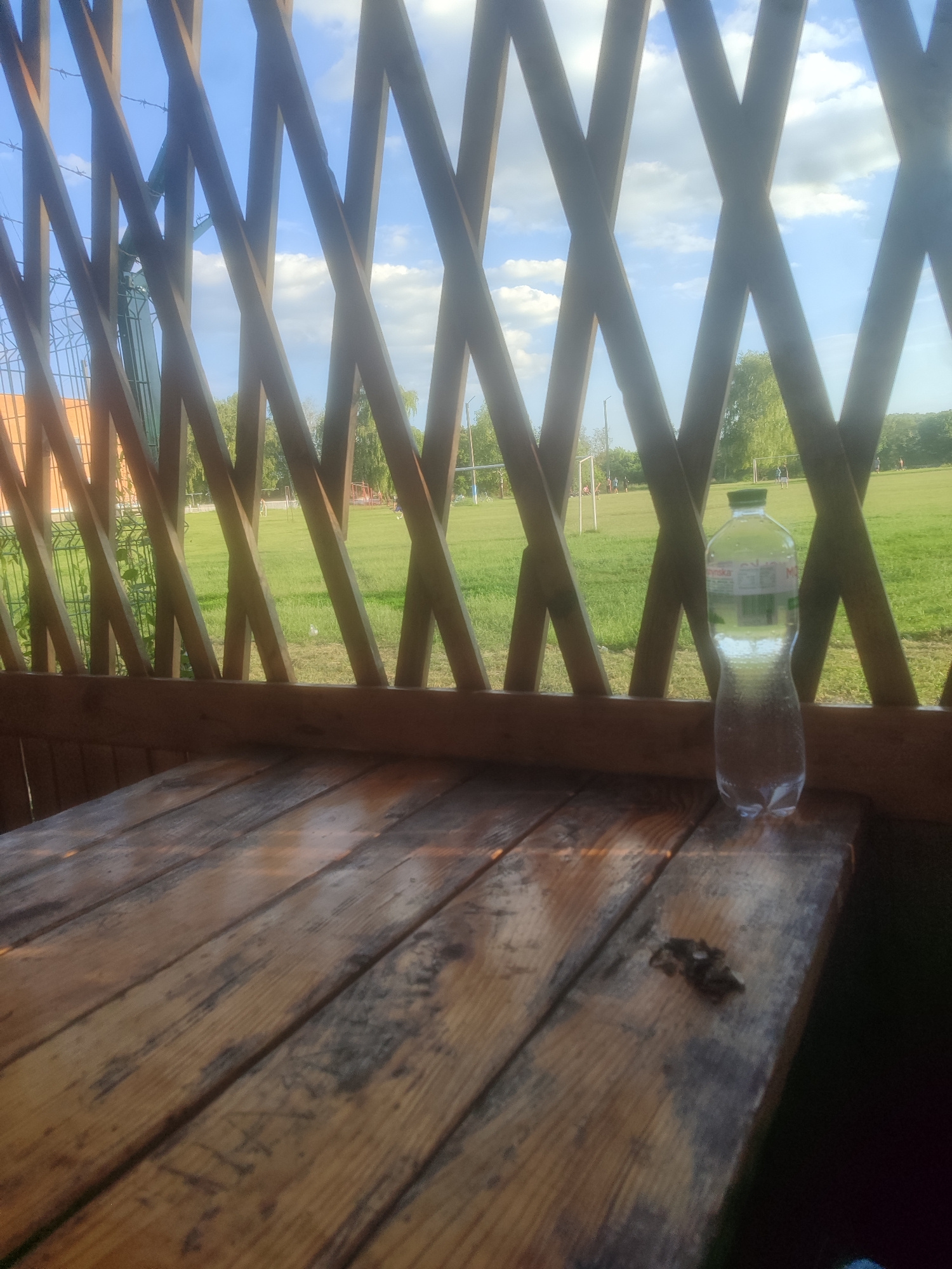 A partially seen wooden table sits in the foreground, viewed from a slightly elevated angle looking towards a grassy field. The table has a weathered appearance with visible grain and dark stains. To the right of the table, a clear plastic bottle of water, partially full, stands upright. Behind the table, a wooden lattice structure creates a barrier, offering a view of the outdoor environment. The background reveals a bright, sunny day with a blue sky dotted with white clouds. A green field is visible, with what appear to be soccer goals and several indistinct figures in the distance, suggesting a park or sports field. Trees line the far edge of the field.