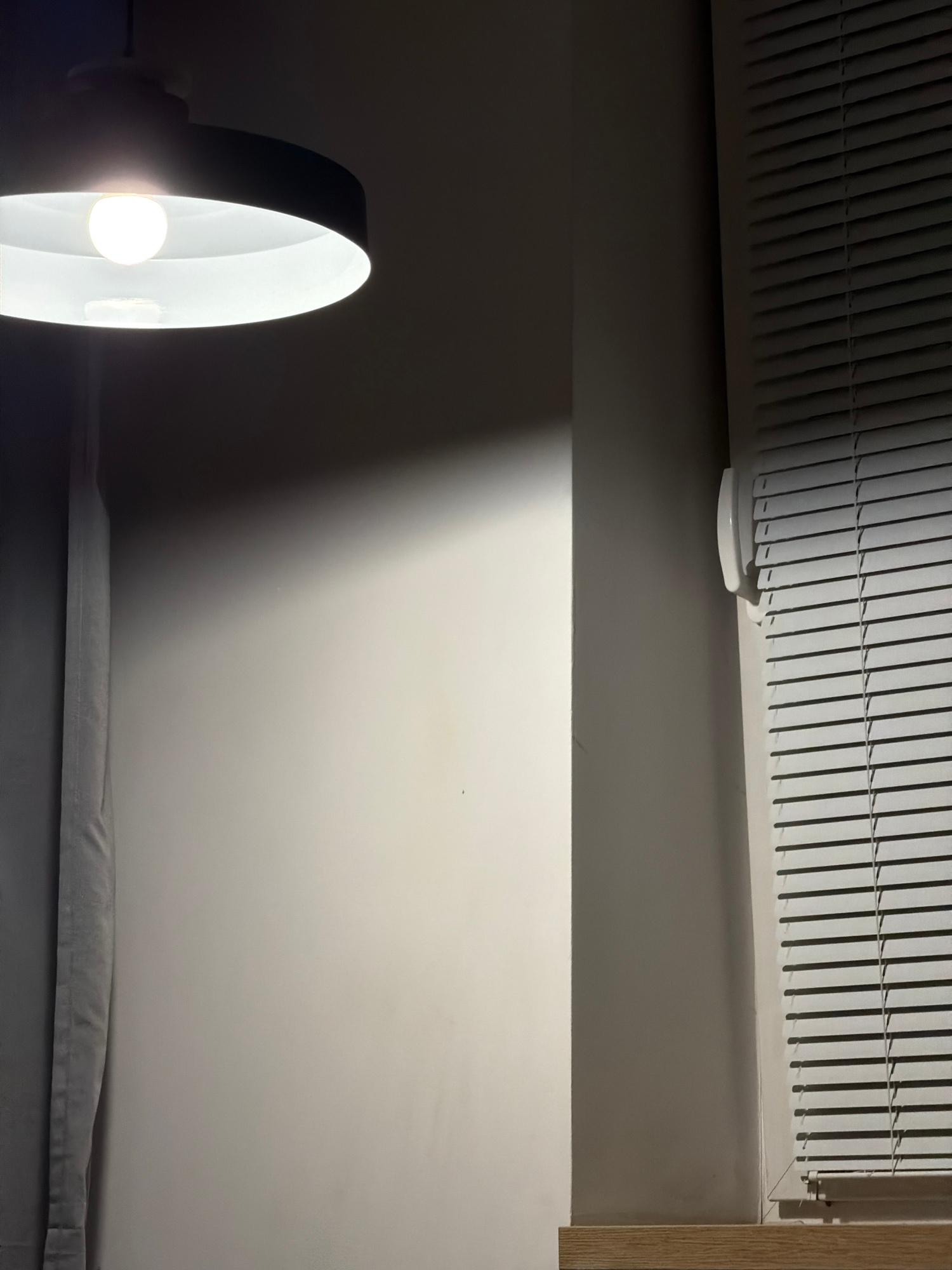 The image depicts an interior scene with a light fixture overhead casting a bright light onto a wall. To the right of the wall, a window with closed blinds is visible. A grey curtain hangs on the left side of the image. The overall lighting suggests an indoor environment, possibly a room in a residential or commercial building. The exact location in Sosnowiec, Poland, cannot be determined from the visual information provided. There are no people, actions, or interactions depicted in the image.