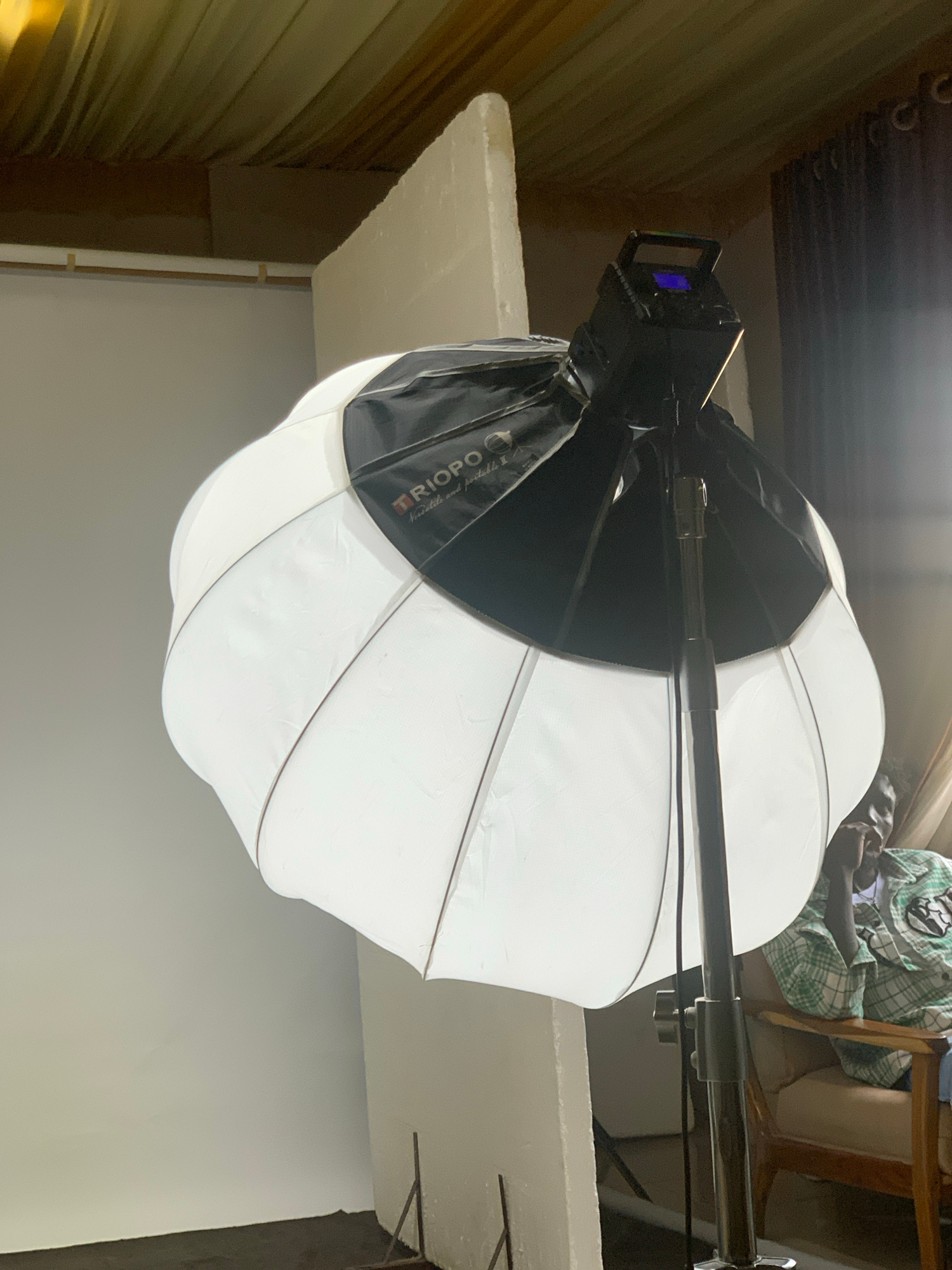 The image shows a studio lighting setup with a large octagonal softbox. The softbox is predominantly white with black accents and appears to be mounted on a stand. A small digital display is visible on the mounting hardware of the softbox, showing a blue screen.
In the background, out of focus, are elements that suggest an indoor setting. There is a corrugated metal wall or ceiling on the left, and a light-colored wall or backdrop on the right. A person is partially visible in the upper right corner, seated in a chair and wearing a green and white checkered garment. The overall lighting is bright, indicating the studio lights are on.
Visible text on the black fabric of the softbox reads "TRIOPO". Below this, there is smaller text that is difficult to fully decipher but appears to be a brand name or model number.
The scene depicts a static setup for photography or videography, with no apparent activity taking place beyond the lighting equipment being set up or in use. The image has a slightly tilted perspective. The context provided indicates this is in Barakin Sabawa, Nigeria.