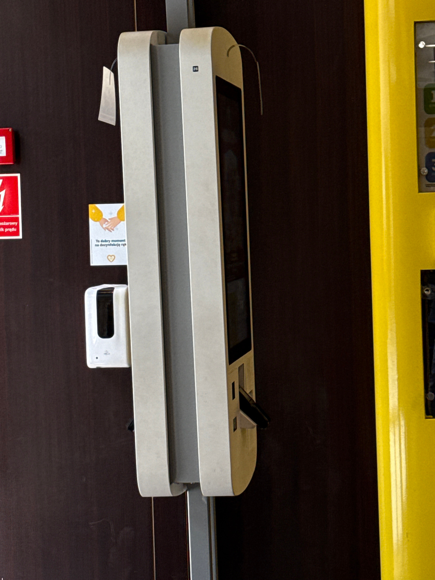The image depicts a close-up, vertically oriented shot of a touch-screen ordering kiosk and a hand sanitizer dispenser mounted on a dark brown wall, possibly in Łódź, Poland, judging by the context provided in the prompt, but this cannot be definitively confirmed from the image itself. The kiosk is a light gray, double-screen unit with a prominent touch display. Below the screen, a card reader is visible. To the left of the kiosk, a white, automatic hand sanitizer dispenser is mounted, accompanied by a small, square informational sign with an illustration of hands being washed and text. A red emergency button is also visible to the far left, partially obscured. The overall environment suggests a commercial setting, possibly a fast-food restaurant or a public service point, where hygiene and ordering are facilitated. The lighting is direct, highlighting the surfaces and the distinct colors of the objects.