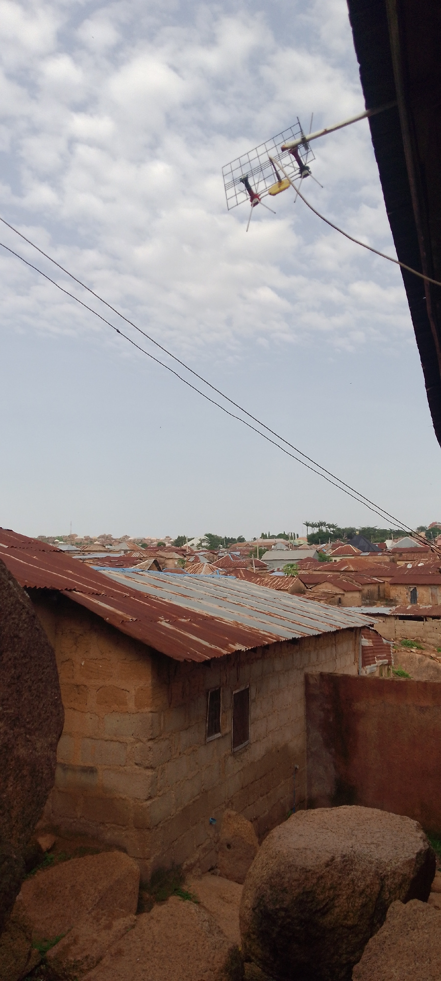 A medium-shot, vertical, outdoor, daylight, eye-level, full-shot image shows a cluster of dwellings with corrugated metal roofs, typical of a developing urban area. In the foreground, a partially visible, light-brown brick building with a rusty, corrugated metal roof is situated among large, rough, brown boulders. The building has two small, dark windows on its visible wall. The background reveals a dense collection of similar structures with predominantly rusty, corrugated metal roofs stretching into the distance. A TV antenna with a white support is mounted on the upper right corner of the frame, extending into the bright, cloudy sky. Two black power lines traverse the sky diagonally from the upper left to the lower right of the image.
