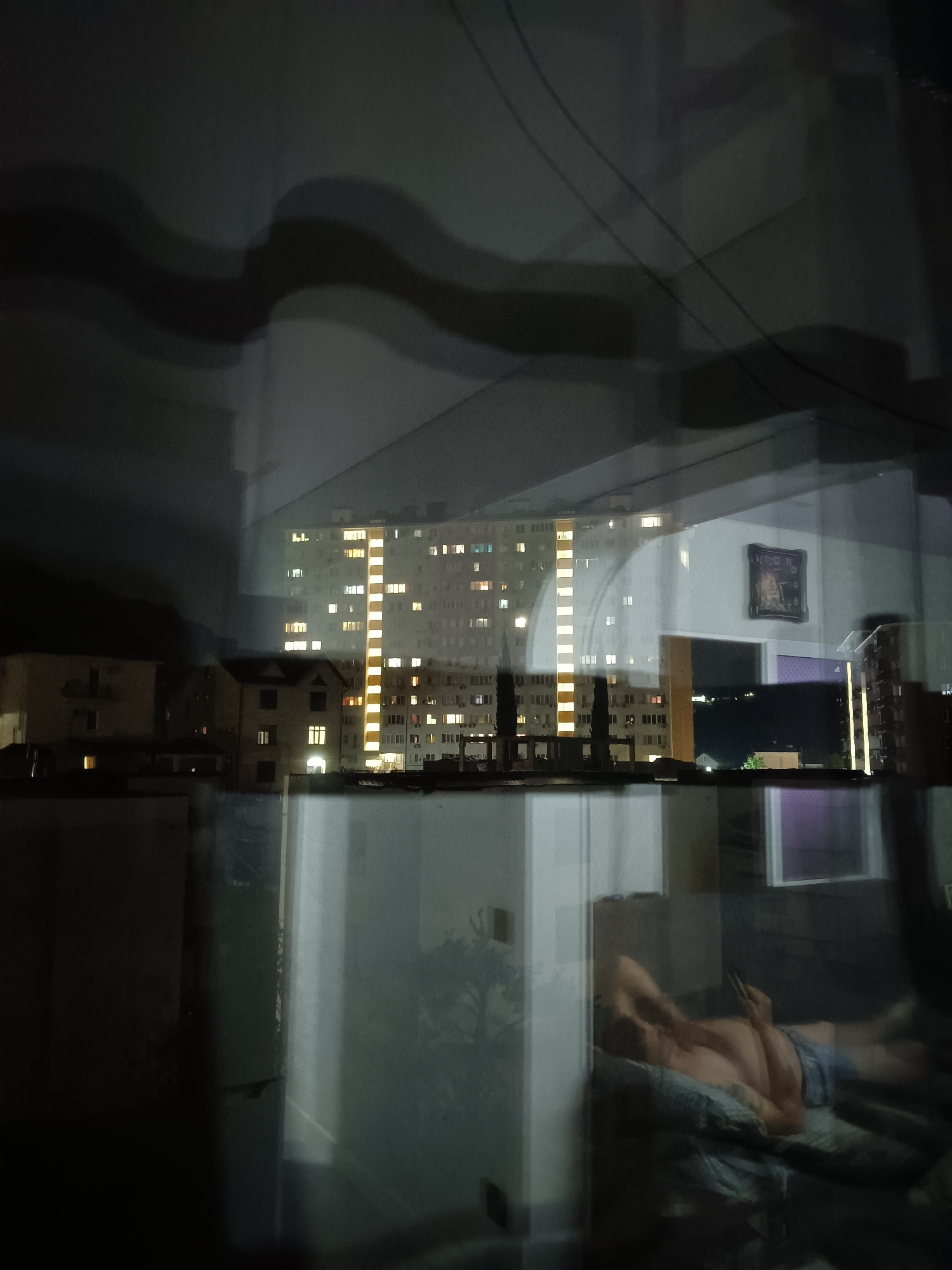 The image captures a night scene through a reflective surface, likely a window. In the background, a brightly lit apartment building dominates the view, its windows illuminated with a warm yellow glow. The building appears to be a multi-story structure, characteristic of urban residential areas. Adjacent to it are other buildings, some of which are also lit.
In the foreground, a person is visible lying down, seemingly on a bed or sofa, illuminated by artificial light from within the room. The person's torso and legs are visible, and they appear to be looking at a handheld device. The reflection on the surface is significant, obscuring parts of the scene and creating a layered effect.
The overall impression is one of an urban environment at night, with the activity within one dwelling reflected against the backdrop of a larger city landscape. The location of Sochi, Russia, is not directly discernible from the visual elements of the image itself; this information would require external context or metadata. However, the architecture of the apartment building, particularly its scale and design, is consistent with residential blocks found in many Russian cities. The presence of power lines or cables can be inferred from thin dark lines visible across the upper portion of the image.