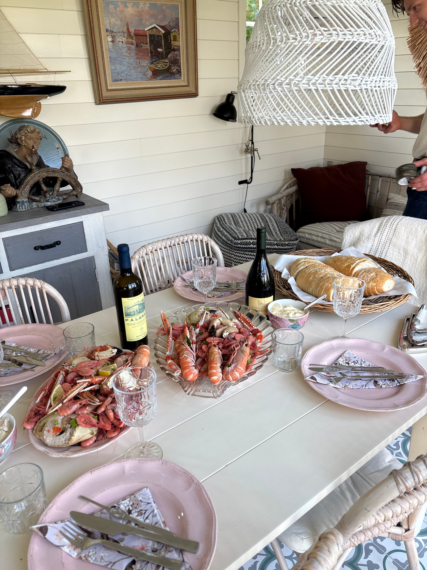 The image depicts an outdoor dining setting on a table laden with seafood, bread, and drinks, likely a terrace or patio. A large white rattan lampshade hangs overhead, and a person's arm is visible on the right, holding a napkin and a silver goblet. The table is set with pink plates, cutlery, and patterned napkins. Several dishes of seafood, including shrimp and langoustines, are prominently displayed. There are also glasses and a bottle of wine. In the background, a painting of a harbor scene hangs on a white paneled wall, and decorative items such as a ship's wheel and a figurine are visible. Wicker chairs surround the table. The floor has a patterned tile design. Based on the provided location of Larkollen, Norway, the scene is situated in this coastal town known for its scenic beauty and maritime activities.