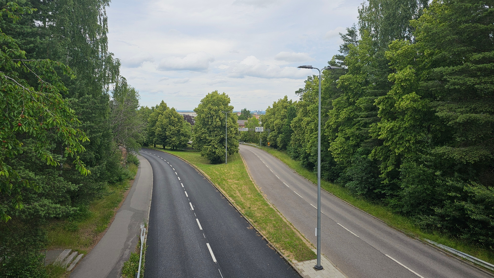 This is a high-angle, eye-level shot of a split highway in Linköping, Sweden. The highway is surrounded by dense, green trees on both sides, with a patch of mown grass in the median separating the two lanes of traffic. The sky is overcast, indicating a cloudy day.
The highway itself is asphalt, with white dashed lines marking the lanes. The left lane is darker, suggesting it might be a newer or more recently paved surface. The right lane is lighter gray. There are streetlights visible along the right side of the road.
In the distance, beyond the trees, hints of buildings and possibly a power line can be seen, suggesting an urban or suburban setting. There are also two road signs visible in the distance, but the text is illegible. The overall scene conveys a sense of calm and everyday transit, with no people or vehicles actively in motion in the frame. The lush greenery and overcast sky suggest it is likely summer or late spring.
