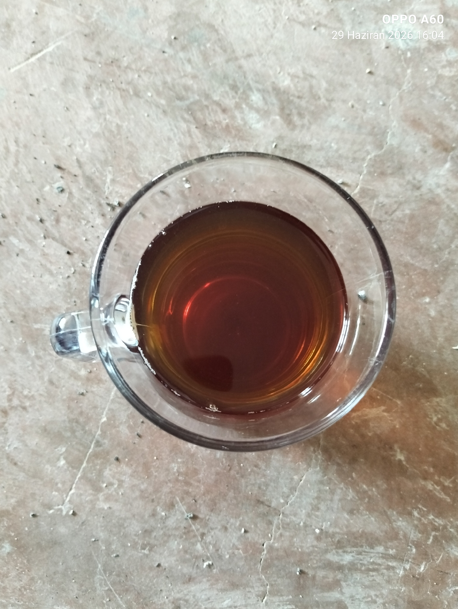 A top-down shot shows a glass mug filled with a dark amber liquid, presumably tea, resting on a textured surface that appears to be a marble countertop. The mug has a clear handle. The surface is speckled with small white and gray particles. In the upper right corner of the image, white text indicates the camera model "OPPO A60" and a date and time stamp "29 Haziran 2026 16:04".