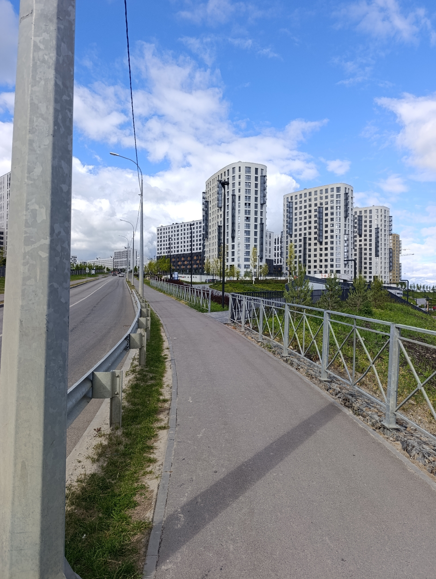 The image shows a modern urban landscape with a clear blue sky dotted with white clouds. In the foreground, a grey metal pole stands to the left, beside a paved road and a pedestrian walkway separated by a metal fence. The road curves to the left, with a guardrail on its right side. Beyond the walkway and fence, a grassy slope rises, dotted with small evergreen trees.
Dominating the background are several tall, multi-story residential buildings. These buildings have a distinct architectural style, featuring curved facades and numerous windows. Some are white, while others appear to have darker accents. The buildings are set against the sky, suggesting they are relatively new constructions. In the distance, more buildings and a construction crane are visible, hinting at ongoing development in the area.
The scene appears to be an outdoor setting, possibly a park or a residential complex. The presence of a road, walkway, and streetlights indicates an inhabited area. The time of day is likely daytime, given the brightness and shadows. The weather seems pleasant, with a mix of sun and clouds. There are no people visible in the frame. The overall impression is one of modern urban development in a suburban or newly developed area. The location is identified as Penza, Russia.