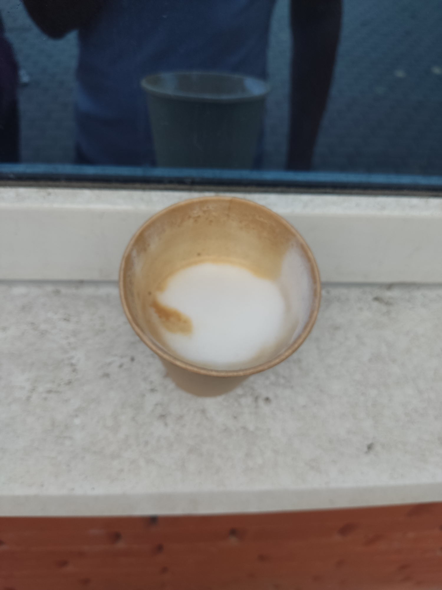 A close-up, overhead shot shows a disposable coffee cup filled with a creamy beverage, possibly latte or cappuccino, resting on a light-colored, textured windowsill. The cup appears to be made of light brown cardboard. The beverage inside has a layer of frothy white foam with a swirl of brown coffee visible beneath. The windowsill is positioned in front of a dark pane of glass, through which a reflection is visible. The reflection shows a person's upper body, including shoulders and arms, wearing a blue shirt, and a dark-colored container, possibly another cup or vase, is also reflected. The background behind the reflection appears to be a blurred outdoor environment. The windowsill is supported by a reddish-brown wooden surface, likely part of a window frame or a piece of furniture. The image is taken in Serpukhov, Russia, indicating an outdoor or indoor setting near a window in that city.