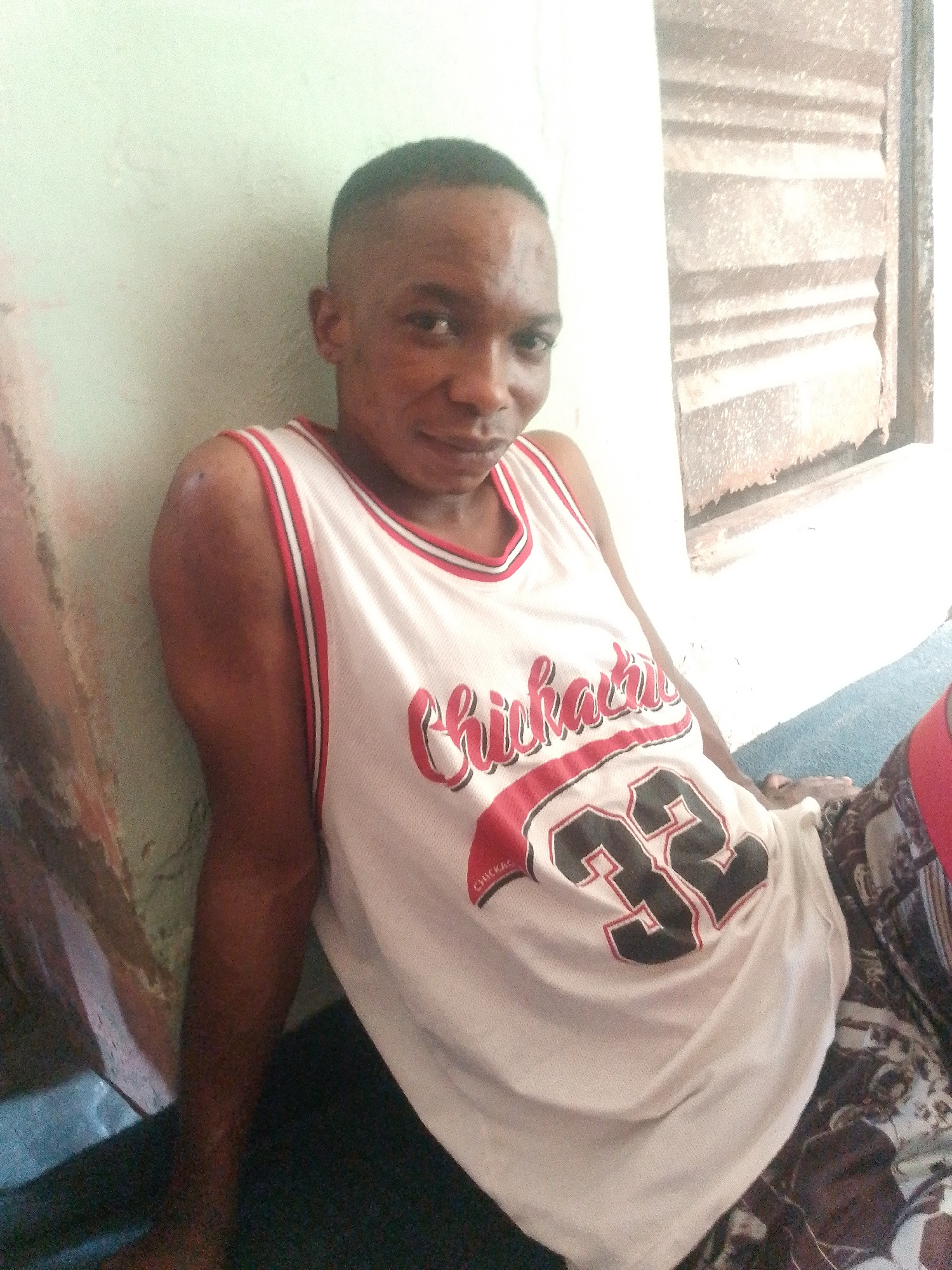 A dark-skinned male sits leaning against a pale green wall with a corrugated metal structure to his right. He is wearing a white basketball jersey with red trim that says "Chicago" in red script and the number "32" in black with a red outline. He appears to be looking at the camera with a slight smile. The lighting is bright and harsh, suggesting daylight. The ground he is sitting on is dark.