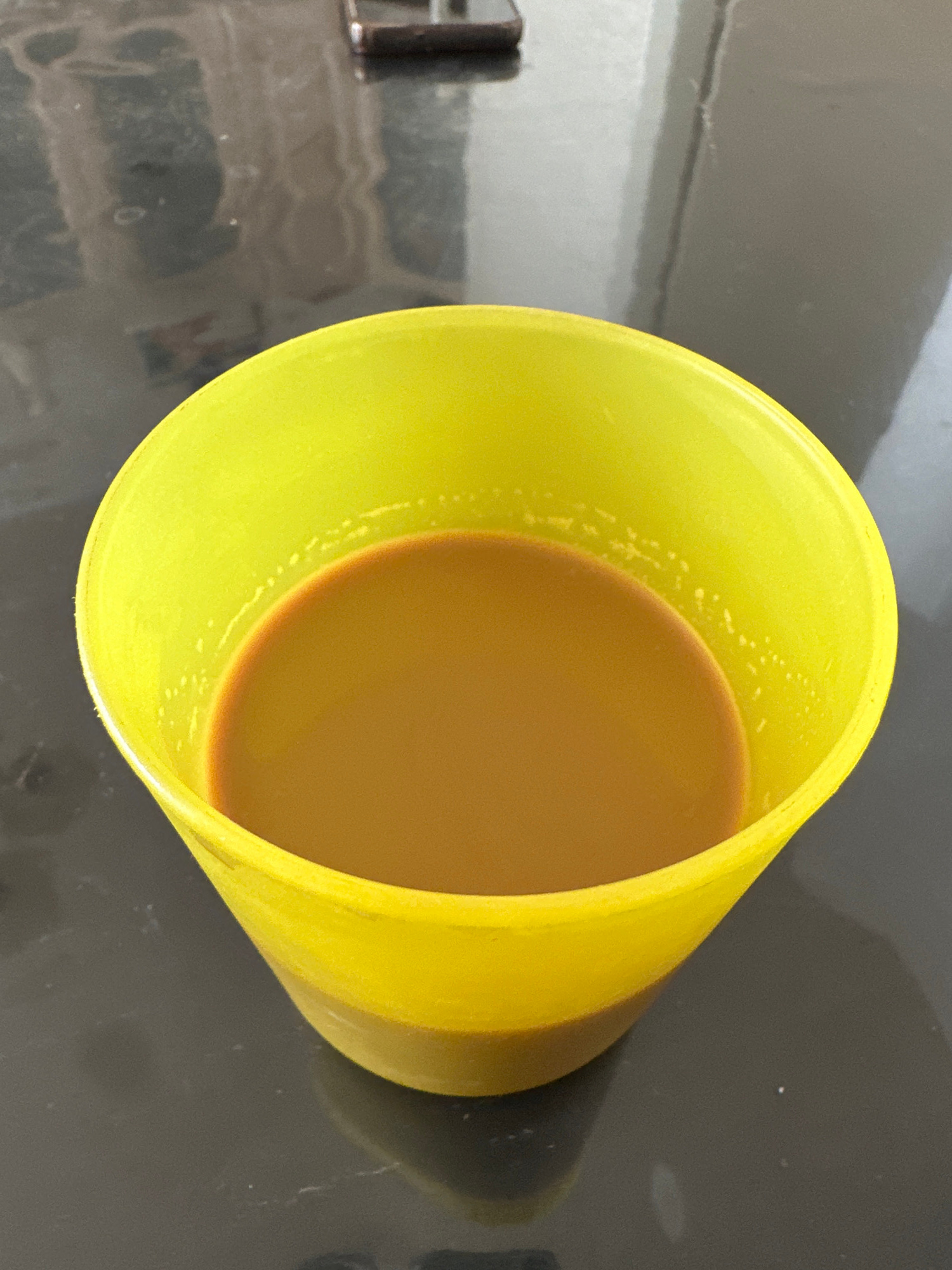 A close-up, high-angle shot shows a bright yellow plastic cup filled with a light brown liquid, likely coffee or tea, sitting on a dark, reflective surface. The liquid's surface is smooth and even, with small bubbles visible along the inner rim of the cup. The background is out of focus, revealing a mottled, gray and white pattern that appears to be a countertop or table with some indistinct objects. The lighting is bright, casting a soft shadow from the cup onto the surface. The overall impression is one of a simple, everyday scene.
