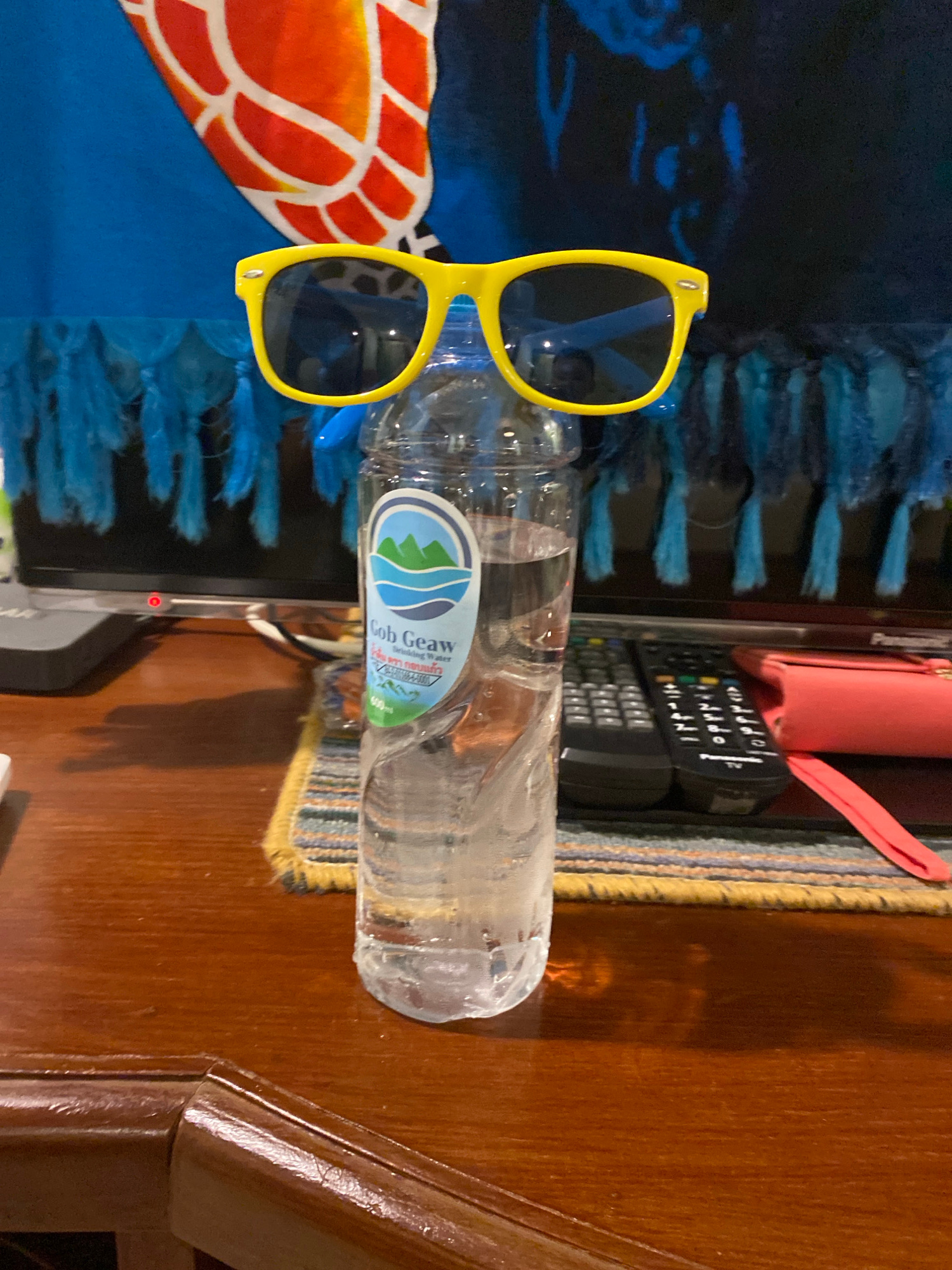 A close-up, eye-level shot captures a plastic bottle of water positioned upright on a wooden surface. Yellow-framed sunglasses, perched atop the bottle, obscure the view of its cap. A portion of a computer monitor, displaying a blue towel with a large orange and white pattern, is visible in the background, along with blue fringe hanging from the towel. Two remote controls and a pink wallet are placed behind the bottle on a patterned placemat. The scene appears to be indoors, possibly in a home or office environment.
