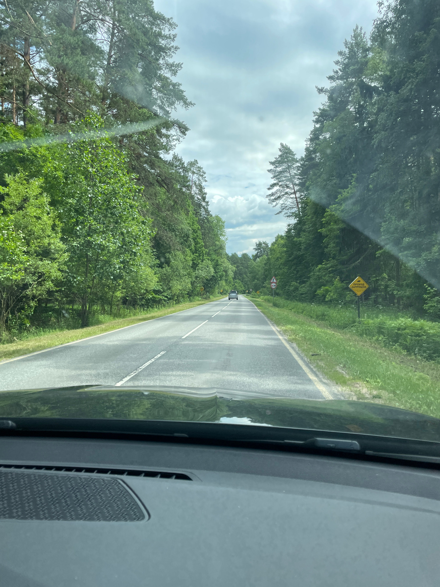 The image captures a view from inside a vehicle, looking forward onto a paved road that winds through a dense forest. The road is asphalt with white lane markings. Lush green trees and foliage line both sides of the road, with a canopy of leaves overhead creating dappled sunlight. The sky is overcast with white clouds. A yellow diamond-shaped sign with black lettering is visible on the right side of the road, and another sign further down the road shows a red circle with a diagonal line through it. Two cars are visible on the road in the distance, one in the center lane and another further ahead. The dashboard of the vehicle is partially visible in the foreground, suggesting the perspective of a driver.