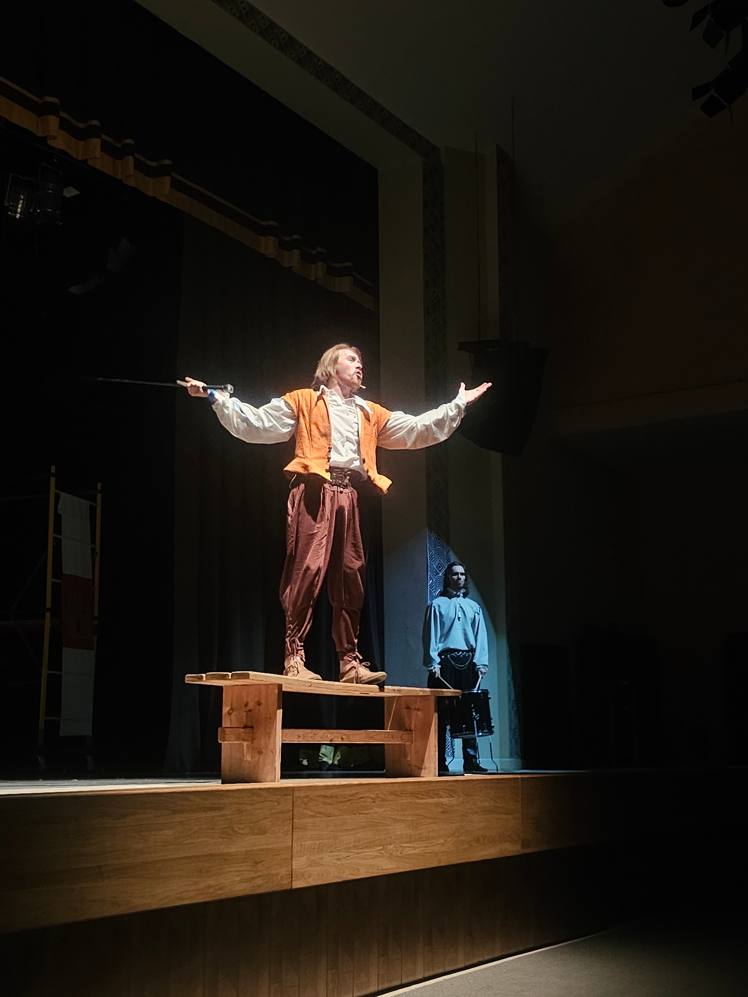 This image captures a theatrical performance on a wooden stage. The main subject, a male performer, stands on a wooden bench, dressed in a white shirt with ruffled sleeves, an orange vest, and brown breeches. He holds a black rod in his outstretched right hand and gestures with his left arm. His hair is light brown and shoulder-length. In the background, another male figure, also in period costume (a light blue tunic and dark pants), stands to the right, holding drumsticks near a black drum. The stage is dimly lit, with spotlights illuminating the central performer. Behind the performers, dark curtains create a backdrop. To the left of the stage, a white and red flag is visible on a yellow structure. The architecture of the venue is suggested by decorative elements on the ceiling and wall. The overall impression is a historical or dramatic play being enacted in an indoor venue. This scene is located in Tosno, Russia, at the Tosno City House of Culture.