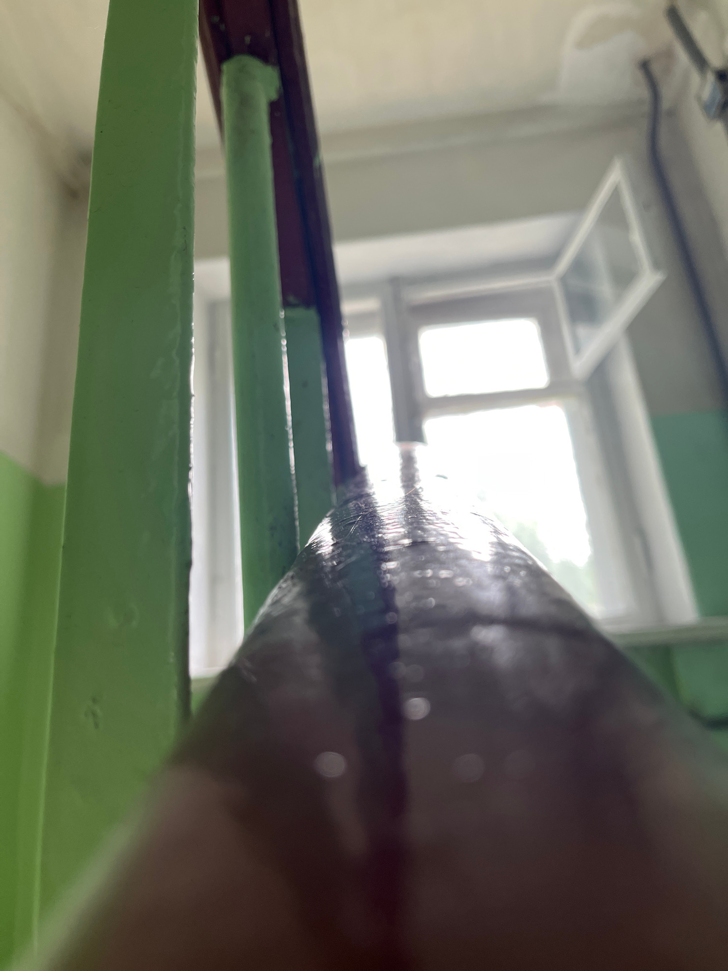 The image is a close-up, low-angle shot of a metallic structure, likely a railing or a part of a staircase, painted in a light green color. The railing appears to be outdoors or in a space with a window. Water droplets are visible on the surface of the railing, suggesting it might be raining or the area is wet.
In the background, a window with white frames is visible. The window has multiple panes and one of the lower panes is open at an angle. Outside the window, it appears to be daytime, with bright light filtering in, possibly indicating overcast or diffused sunlight. The surrounding walls seem to be a light color, possibly white or cream. A dark, possibly brown, cylindrical object is in the foreground, partially obscuring the view of the railing and the window.
The scene is likely set in a building, possibly an apartment complex or a public space. The presence of the railing and the window suggests an interior space overlooking an exterior, or a balcony. The lighting is bright but diffused, making it difficult to determine the exact time of day, but it appears to be daytime. There are no people visible in the frame. The overall impression is one of a mundane, everyday setting, possibly with some recent precipitation. There is no visible text in the image.