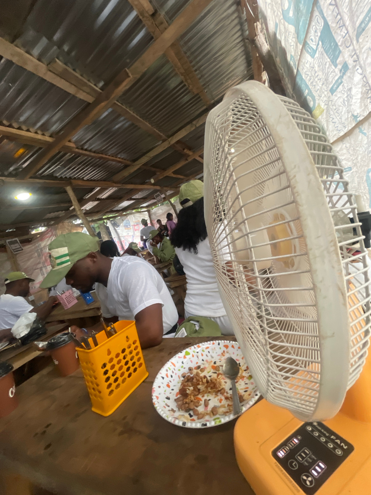 The image depicts an indoor setting, likely a makeshift eatery or gathering space, with a corrugated metal roof supported by wooden beams. A white oscillating fan stands prominently in the foreground on the right, its blades partially visible. The fan is mounted on an orange base with control buttons and indicator lights.
In the midground and background, several individuals are seated at wooden tables, some wearing light-colored t-shirts and green caps with insignia, suggesting a possible uniform or group affiliation, such as the National Youth Service Corps (NYSC) program common in Nigeria. Some individuals appear to be eating. On the tables, there are plates of food, cutlery, and containers. One plate in the foreground contains rice and beans. An orange plastic utensil holder is also visible on a table.
The environment appears to be rustic and informal, with visible construction materials and possibly makeshift walls or coverings in the background. The lighting is somewhat dim, with a bright light source visible in the upper left portion of the image. The scene appears to be set in Orun Ekiti, Nigeria, given the context provided.