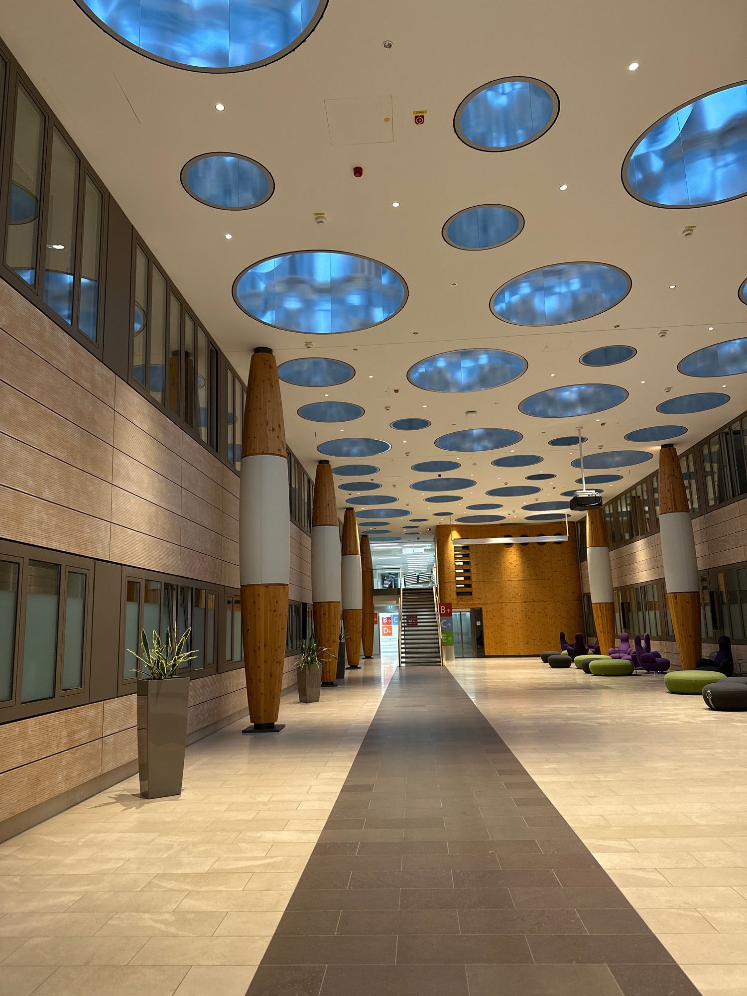 The image depicts an interior hallway with a distinctive ceiling featuring numerous circular, blue-lit skylights. The hallway is flanked by walls with horizontal wooden paneling and large windows. Several large, cylindrical pillars with a wooden texture and white sections run along the hallway. Planters with green foliage are positioned near the pillars.
In the distance, a staircase is visible, leading to an area with wooden paneled walls. The floor is tiled with large, light-colored stone tiles. To the right side of the hallway, there are clusters of colorful, modern seating arrangements, including purple and green, bean-bag-like chairs, and darker, circular poufs. Some of these seating areas are occupied by dark silhouettes, suggesting the presence of people, though their features are not discernible. The overall environment appears to be a modern public space, possibly an office building, a convention center, or a public institution.
This image was taken at the Bavarian Center for Applied Energy Research (ZAE Bayern) in Garmisch-Partenkirchen, Germany.