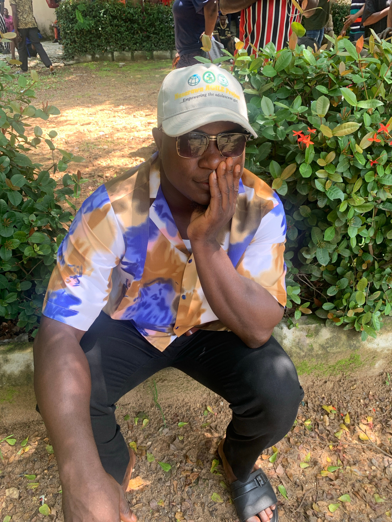 A man wearing a light-colored baseball cap with logos, sunglasses, and a patterned short-sleeved shirt sits in a squatting position on the ground. He is resting his chin on his hand, and his other arm is bent with his elbow on his knee. The background consists of green foliage, dirt, and a concrete barrier. The man is wearing black pants and black slides. The image appears to have been taken outdoors in a natural setting, possibly in Akwanga, Nigeria, given the context. Other people are visible in the background, some walking and others appearing to be engaged in an activity. The lighting suggests it is daytime.