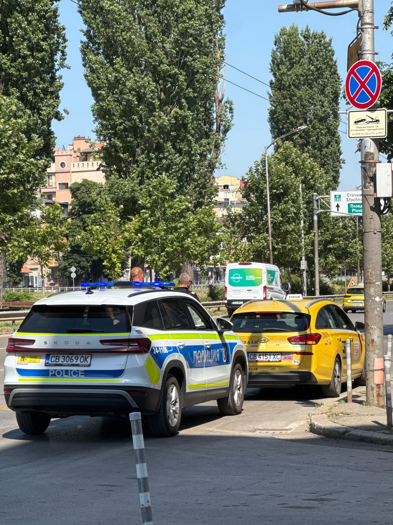 A police Skoda Kodiaq and a yellow taxi are parked on a city street in Sofia, Bulgaria, with tall green trees and buildings in the background. The police car, with its blue lights illuminated, is labeled "POLICE" in English and Bulgarian. The yellow taxi has the word "TAXI" on its roof. A traffic sign indicating "No Stopping" is visible on a pole, along with a sign indicating directions to "Stoechna" and "Plovd". The sky is clear and blue, suggesting a sunny day.
