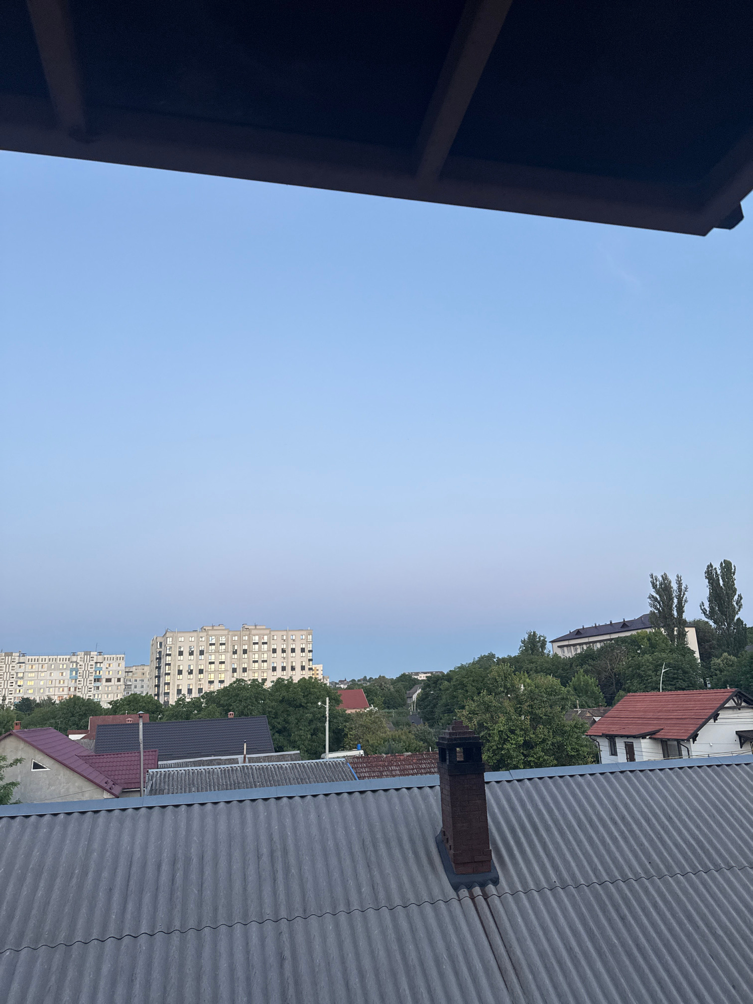 The image captures an outdoor scene at an elevated viewpoint, likely a balcony or rooftop, looking out over a cityscape. The foreground is dominated by the corrugated texture of a grey roof and a brick chimney. Beyond this, a cluster of residential buildings with red and grey roofs are visible, interspersed with green trees. Further in the background, taller, modern apartment buildings rise against a pale blue sky, indicating an urban environment. The overall lighting suggests either early morning or late evening.