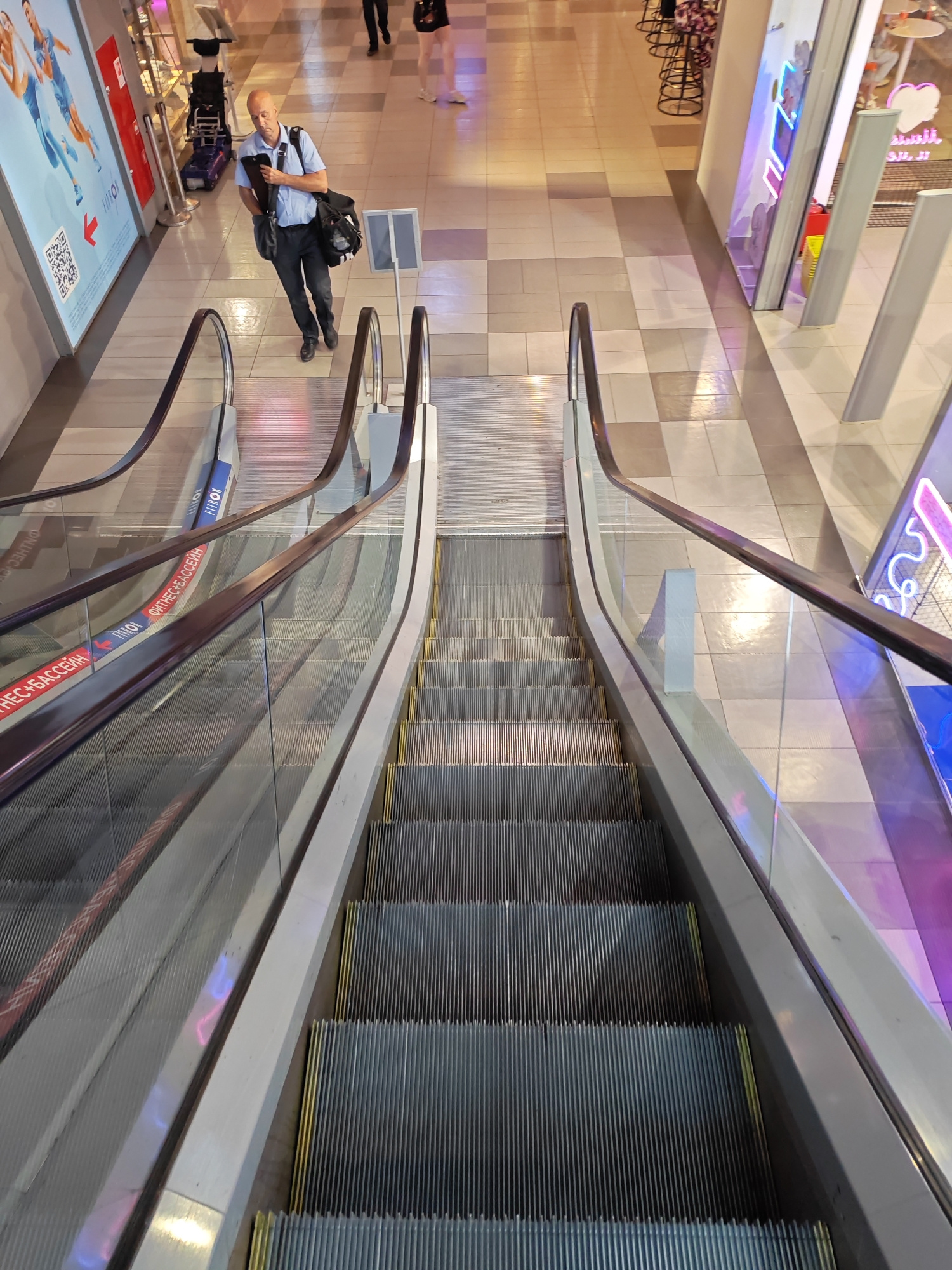 An overhead, high-angle shot shows two parallel escalators moving downwards inside a brightly lit shopping mall. On the left escalator, a bald white man in a light blue short-sleeved shirt, dark pants, and black shoes is walking down. He is carrying a black shoulder bag and a black folder under his left arm. He is looking downwards. Several other shoppers are visible in the background, also moving downwards on the escalators or walking on the tiled floor. Advertisements and shop windows with neon signs are visible on the sides of the escalators. The floor is tiled with alternating light and dark square tiles. This scene is located in Rostov-on-Don, Russia, specifically inside the "Platov" shopping and entertainment center, as indicated by the visible text on the escalator.