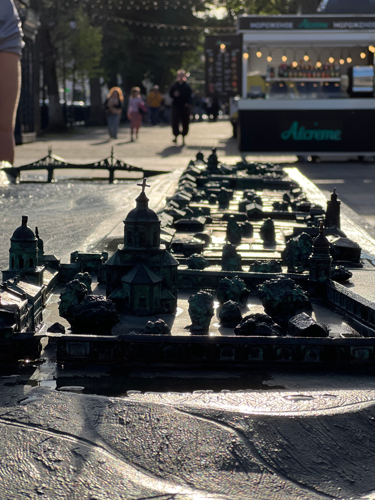 The image shows a sunlit, miniature architectural model, likely representing buildings and possibly a cityscape. The model is made of a dark, metallic material, possibly bronze or weathered metal, and is detailed with small structures, including domes and spires, one topped with a cross. The sun glints off the surfaces of the model and catches water droplets, suggesting it might be part of a fountain or has recently been rained on. In the background, out of focus, are people walking along a paved area. Further in the background, a large dark sign with bright lights and green text is visible, reading "Aliviemme". The overall impression is of an outdoor public space, possibly a park or a pedestrian area, illuminated by bright sunlight, indicating a clear day. The time of day appears to be late afternoon, given the long shadows and the angle of the sun.