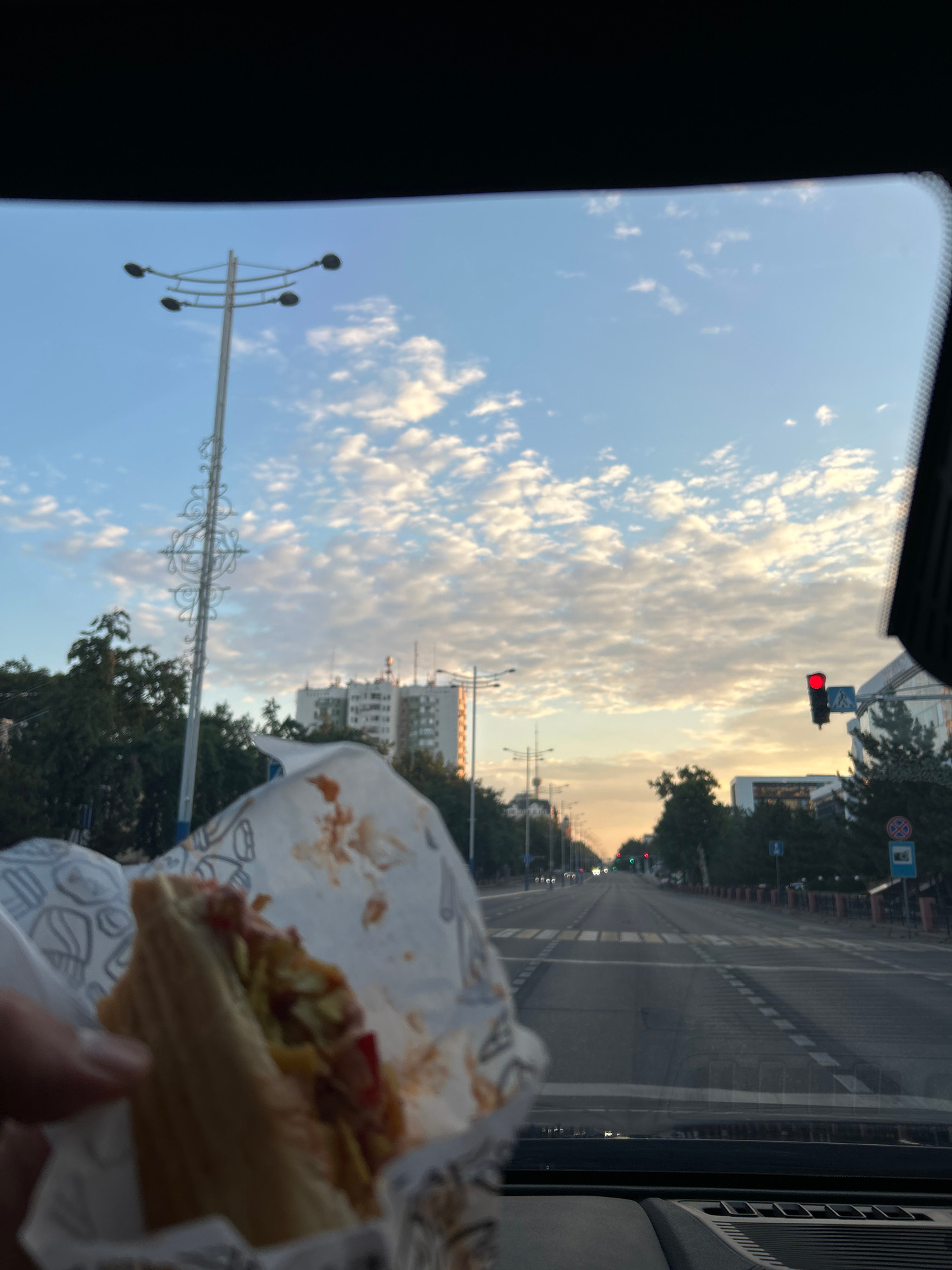 The image shows a view from inside a vehicle, looking through the windshield. In the foreground, a person's hand is holding a sandwich wrapped in white paper, with some red sauce and lettuce visible. The background depicts a wide, empty road with multiple lanes, lined with trees and buildings. The sky is blue with scattered white clouds, and the sun appears to be setting, casting a warm glow on the horizon. A traffic light is visible, showing a red signal. Streetlights and power lines are also present. The scene is set in Tashkent, Uzbekistan, on Amir Timur Avenue, as evidenced by the distinctive lampposts and the architectural style of the buildings.