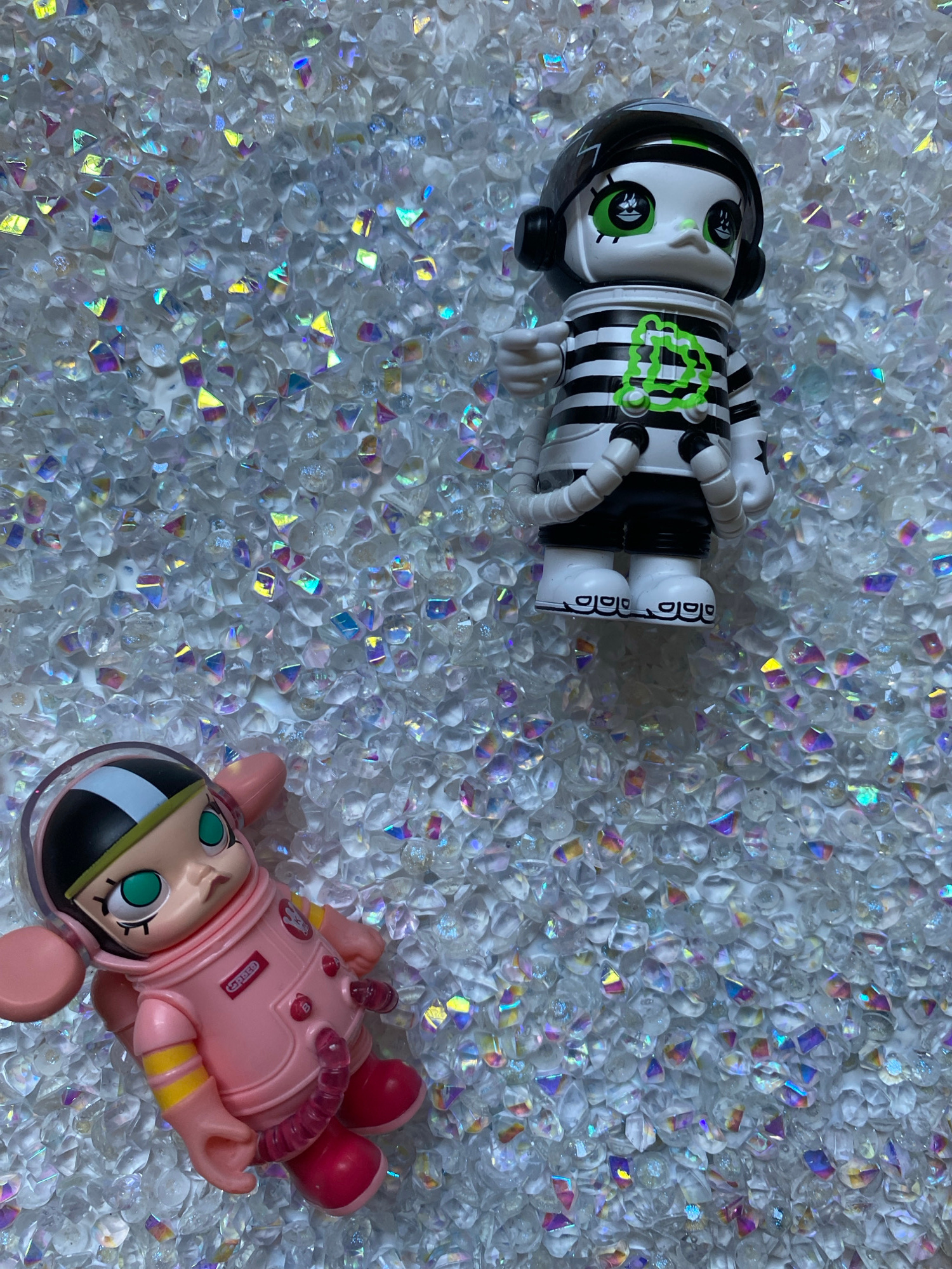 Two collectible figurines are displayed on a surface covered with small, clear, faceted crystals that exhibit iridescent refractions of light. In the upper right portion of the image is a figurine of a character wearing a black helmet with a green stripe, headphones, and a black and white striped shirt with a green neon symbol on the chest. It has white skin with green eyes and appears to be a bird-like creature. In the lower left portion of the image is a pink figurine wearing a pink astronaut suit with yellow accents and a clear helmet. This figurine has large green eyes and appears to be lying down. The background is a dense layer of the iridescent crystals. The image does not depict any specific location in Chernivtsi, Ukraine, as it is an indoor still-life composition.