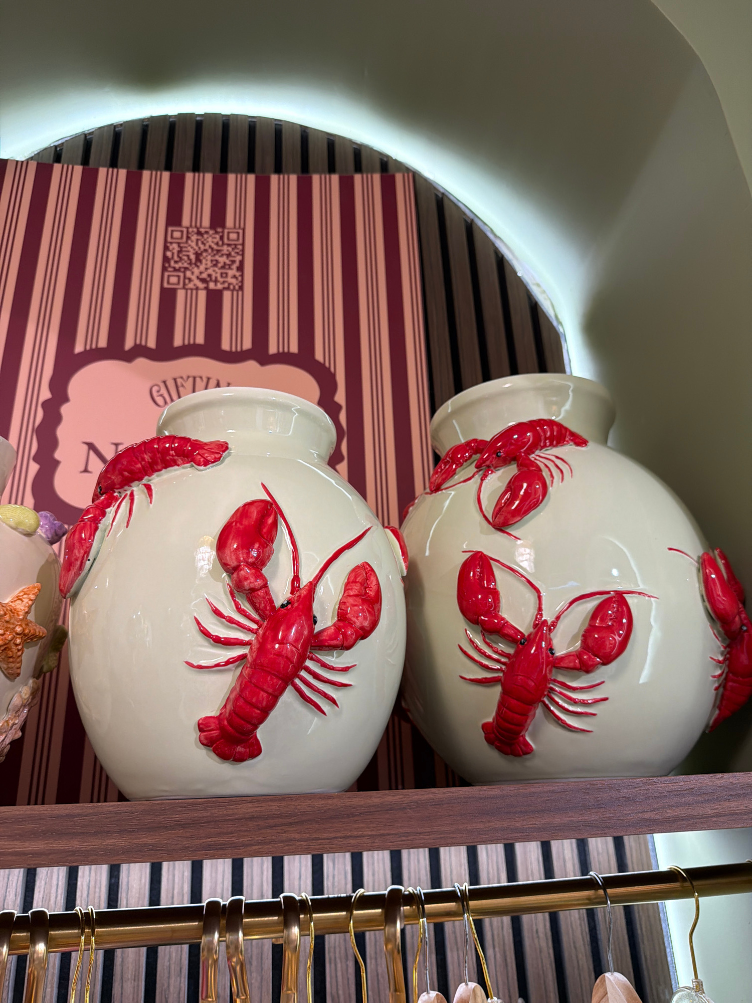 Two cream-colored ceramic vases, each adorned with several raised red lobsters, are displayed on a wooden shelf. Behind the vases, a striped background in shades of burgundy and cream is visible. The vase on the left also features a starfish and seashells. The shelf is situated above a row of golden hangers. The scene appears to be an indoor retail display, possibly in Mdina, Malta, given the context of the request.