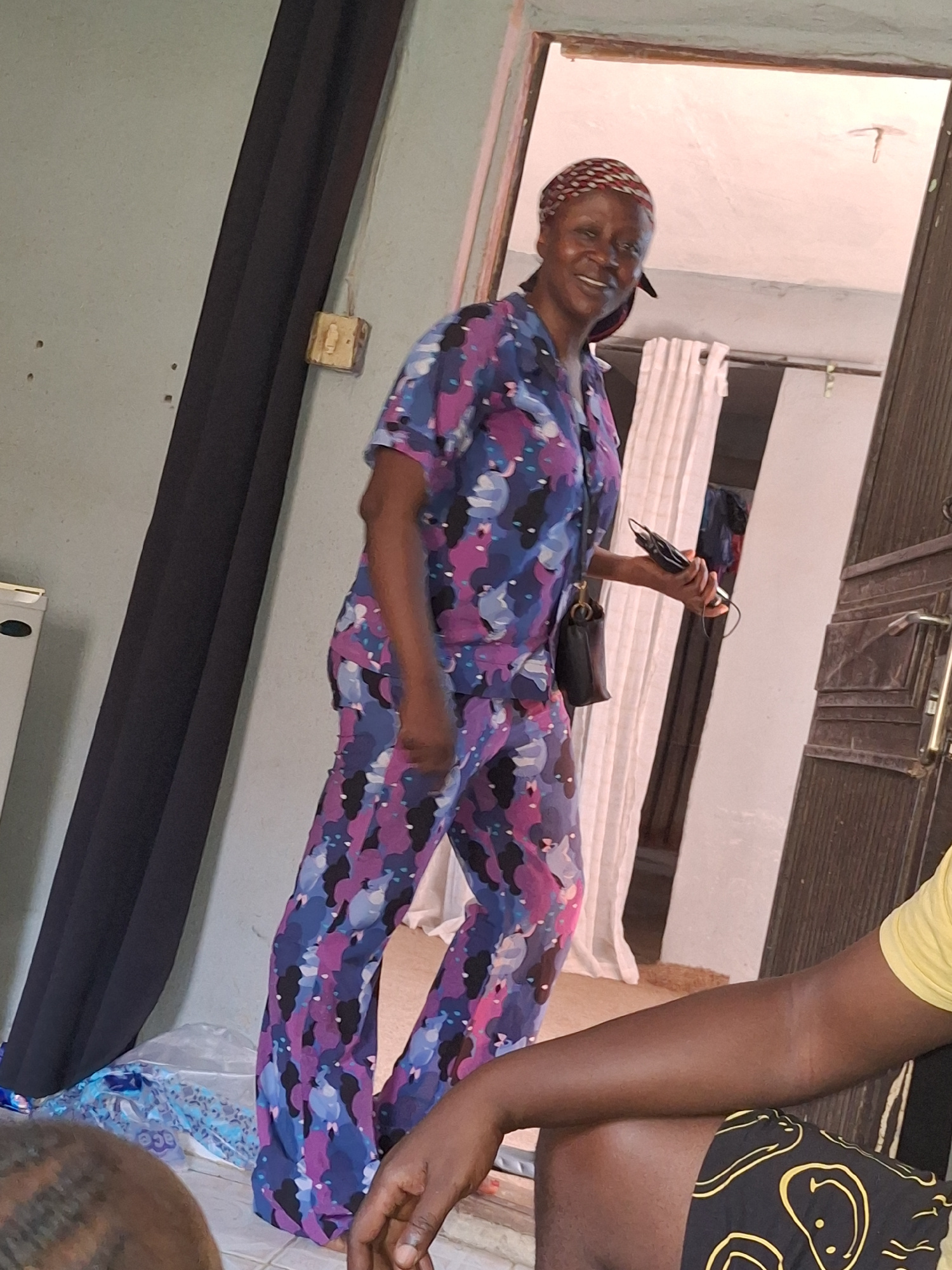 A medium shot captures a Black woman from the knees up, standing indoors and smiling at the camera. She is wearing a colorful, patterned pajama set in shades of blue, purple, and pink, and a headscarf. Her right hand holds a phone. To her left, a black curtain hangs vertically. To her right, a brown wooden door and a white wall are visible. A portion of another person's arm, wearing a yellow shirt and patterned shorts, is in the foreground on the right. The woman is positioned near an open doorway that leads to another room, where a white curtain is visible. The lighting is natural and seems to be coming from the open doorway. The overall scene appears to be a domestic interior in Sabon Tasha, Nigeria.