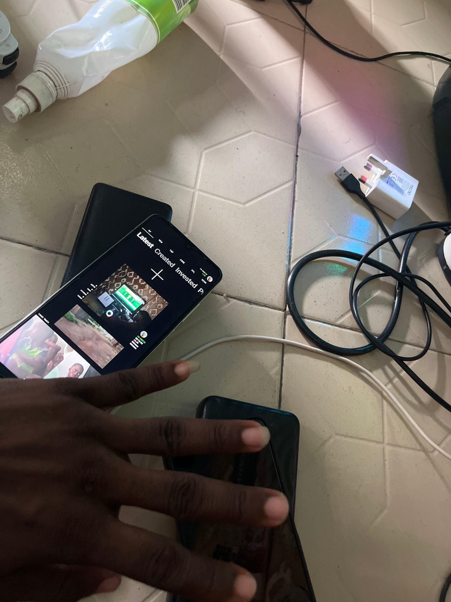 A dark-skinned hand with light-colored fingernails is visible in the foreground, positioned over two smartphones lying on a light-colored tiled floor. The smartphone on top displays a dark screen with some video thumbnails, while the smartphone underneath is black and partially obscured. Various charging cables and a white wall charger with a USB port are scattered around the phones, along with a crumpled white bottle with a green label. The lighting suggests an indoor environment, possibly a room, with shadows cast on the floor.